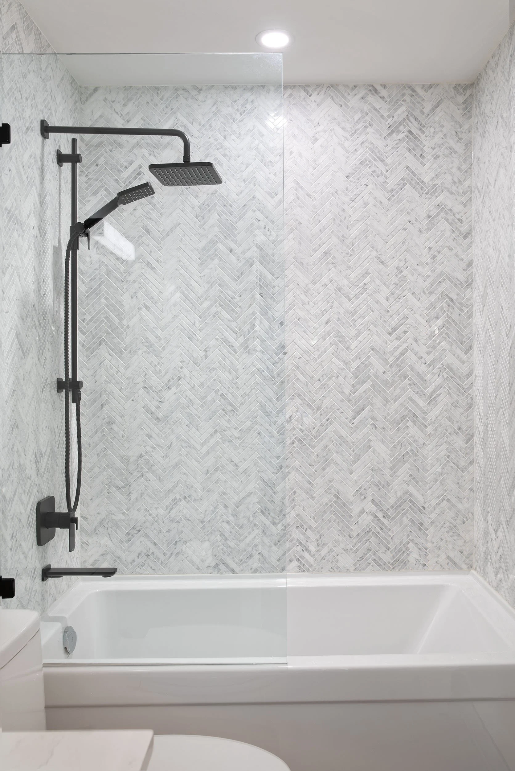 Bathroom shower area with black wall-mounted adjustable shower head, a square rain shower head, a handheld shower, and a glass shower door, with marble-patterned tiles on the walls.