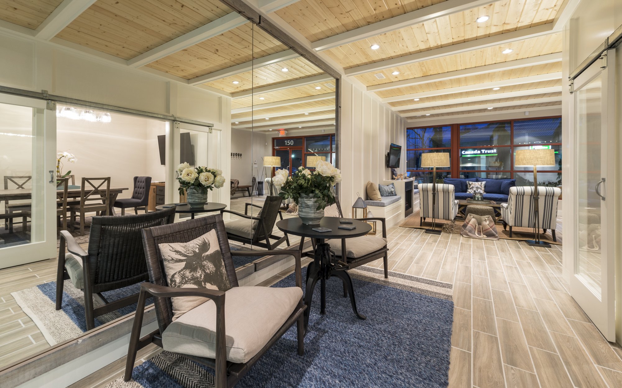 Interior of a modern living and dining area with wood-paneled ceiling, white walls, and large windows. The space is decorated with plants, lamps, and comfortable seating including a sofa and armchairs.