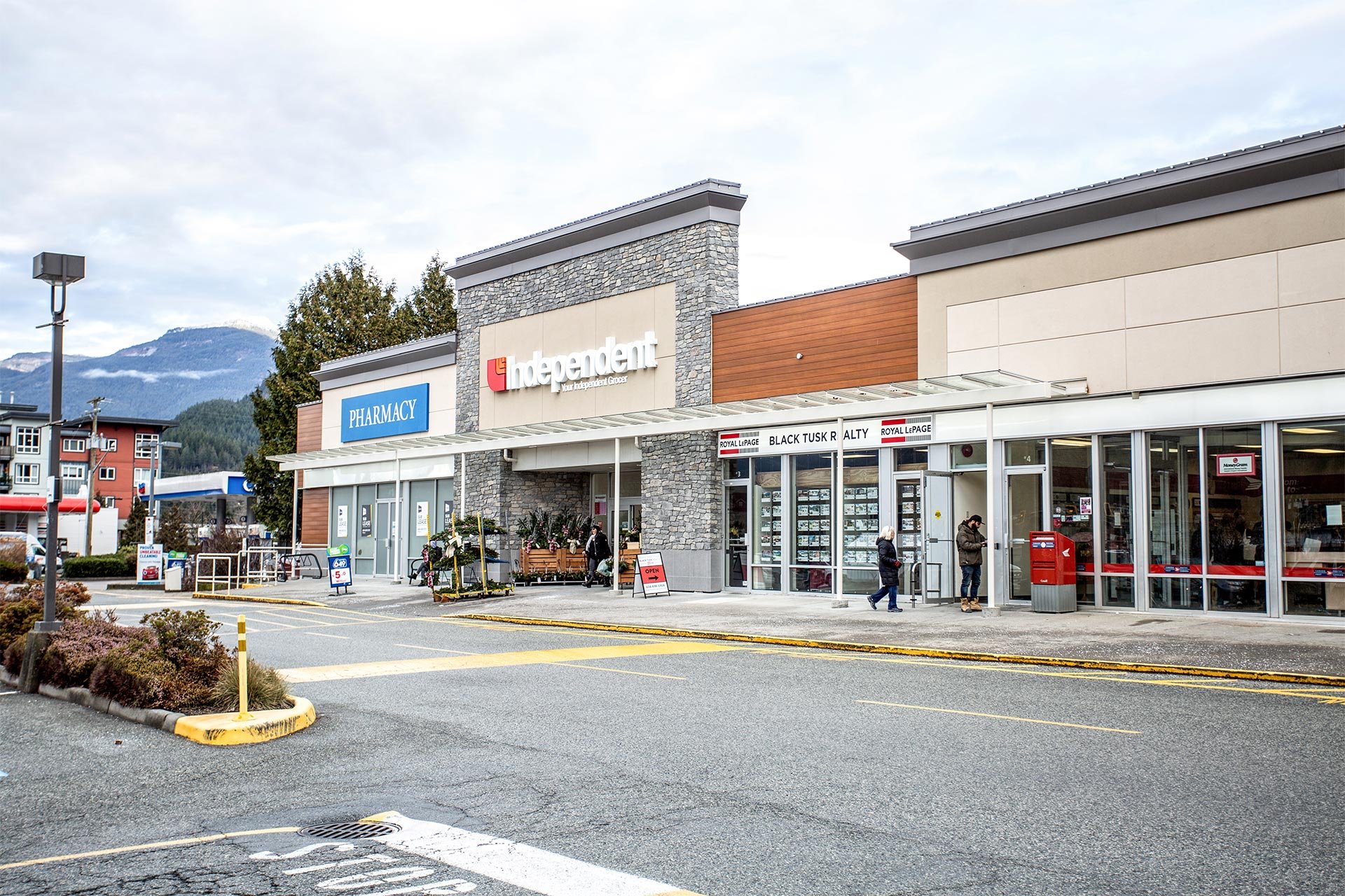 A retail shopping center with a pharmacy, an independent store, and other storefronts in a suburban area, with a mountain backdrop and a cloudy sky.