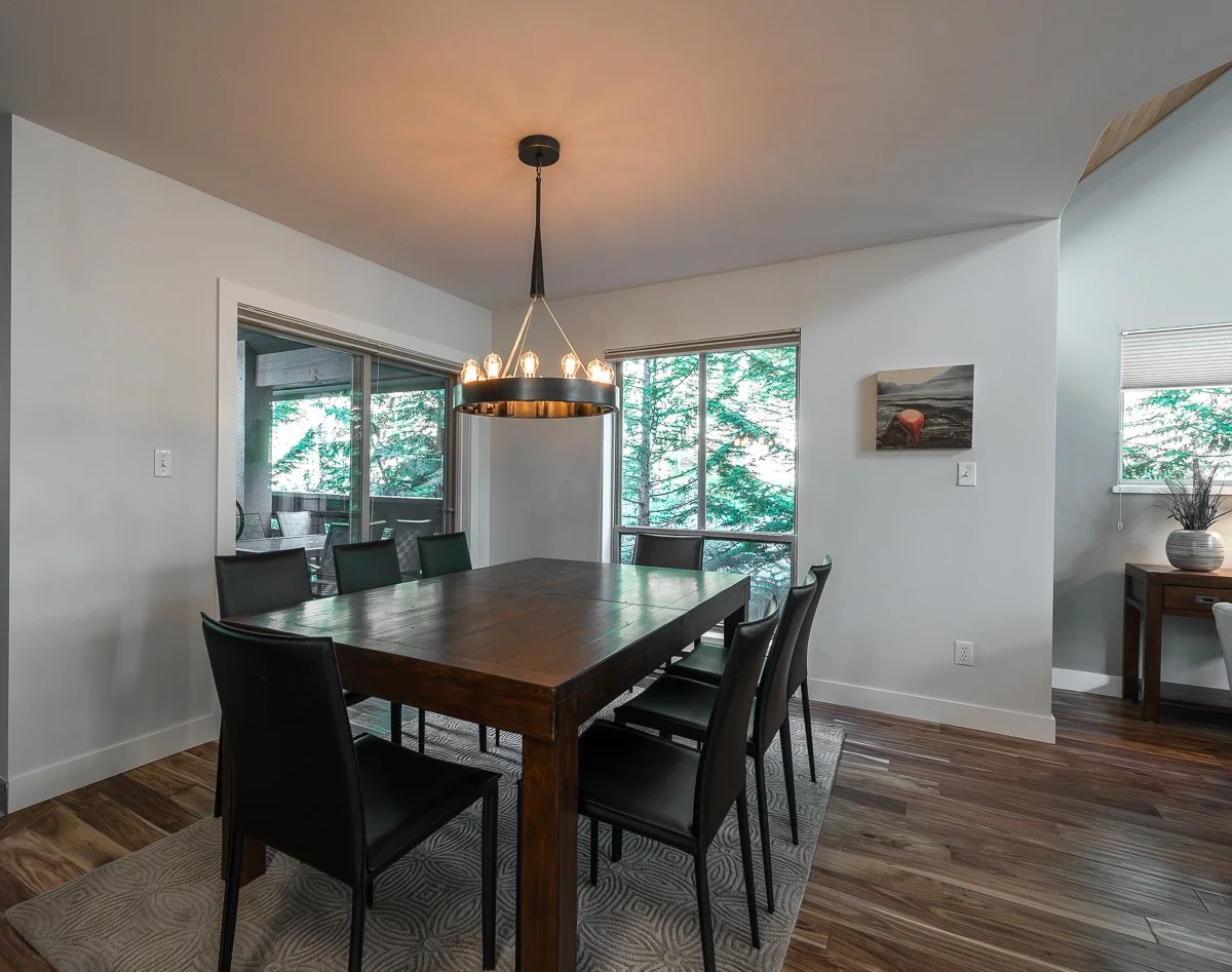 Modern dining room with a wooden table, black chairs, a chandelier, and large windows showing trees outside.