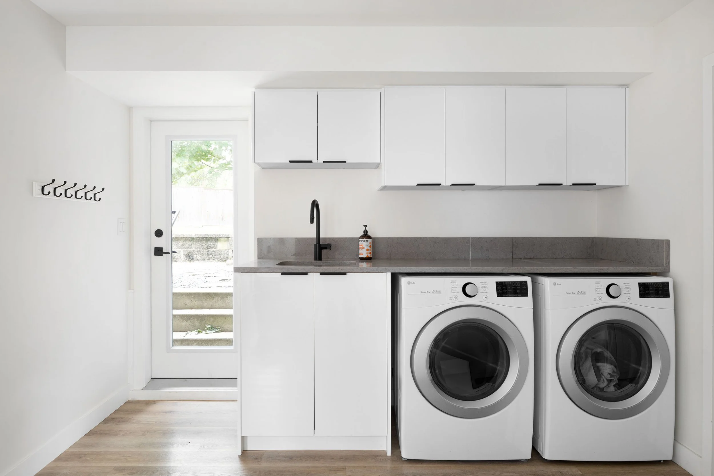 A laundry room with white cabinets, a black sink faucet, a bottle of soap, and a washing machine and dryer side by side. A door with a window leads outside, and a wall-mounted coat hook is on the left.