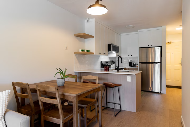 Kitchen and dining area in an apartment with a wooden dining table, four chairs, a small plant, white cabinets, a stainless steel fridge, and a yellow door in the background.