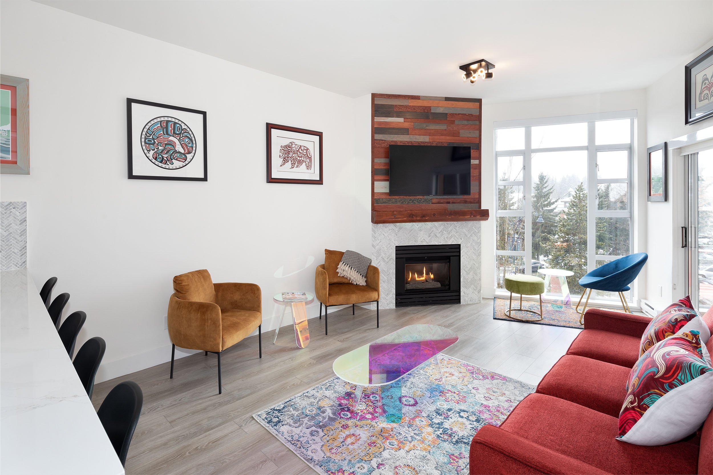 A modern living room with a fireplace, a wall-mounted TV, art on the walls, and bright large windows showing snowy outdoors. There are colorful chairs and a rug on the floor.