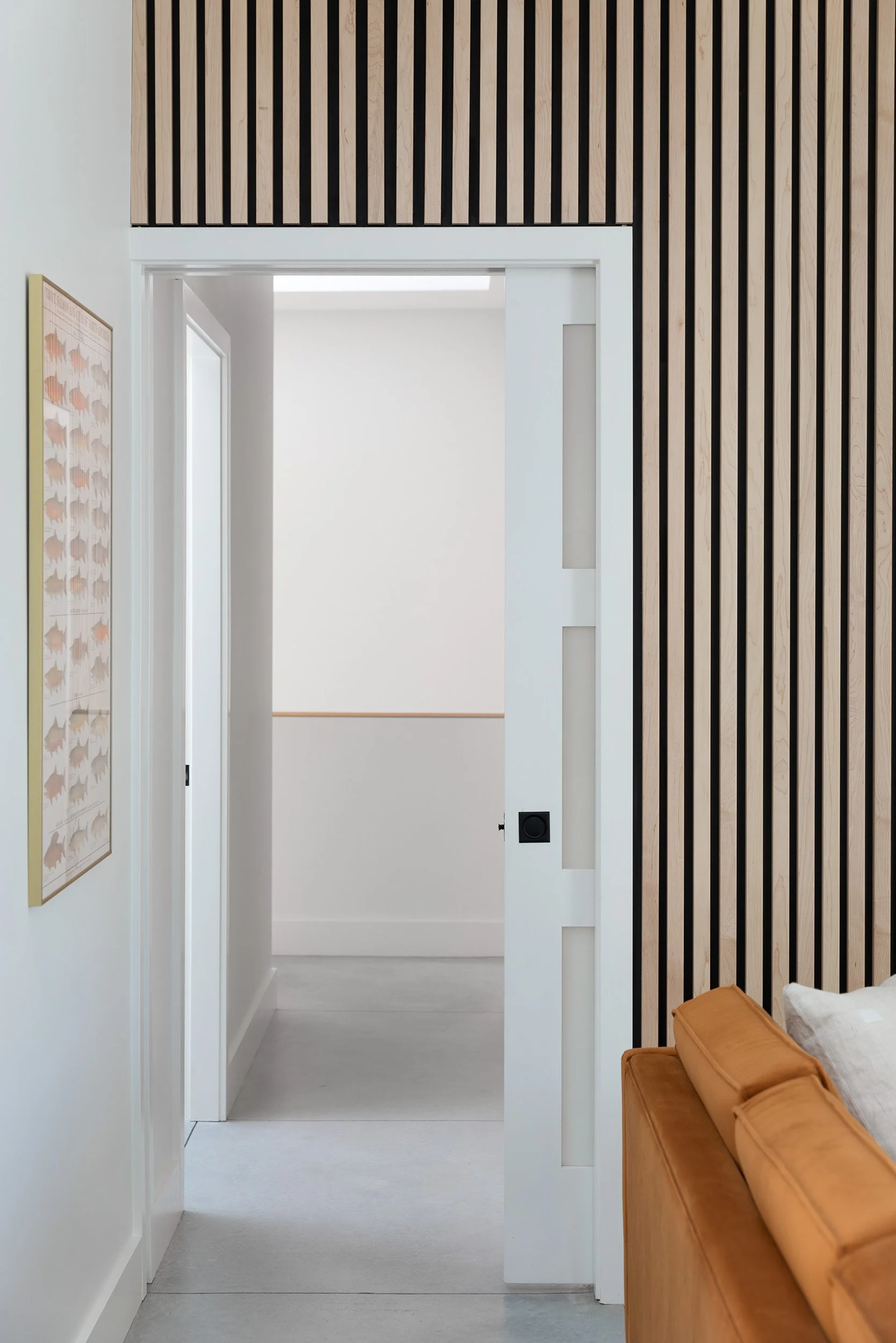 A hallway with white walls and a white door frame leading to another room. On the right, there's a wall with vertical wooden slats. Part of an orange sofa is visible in the bottom right corner.