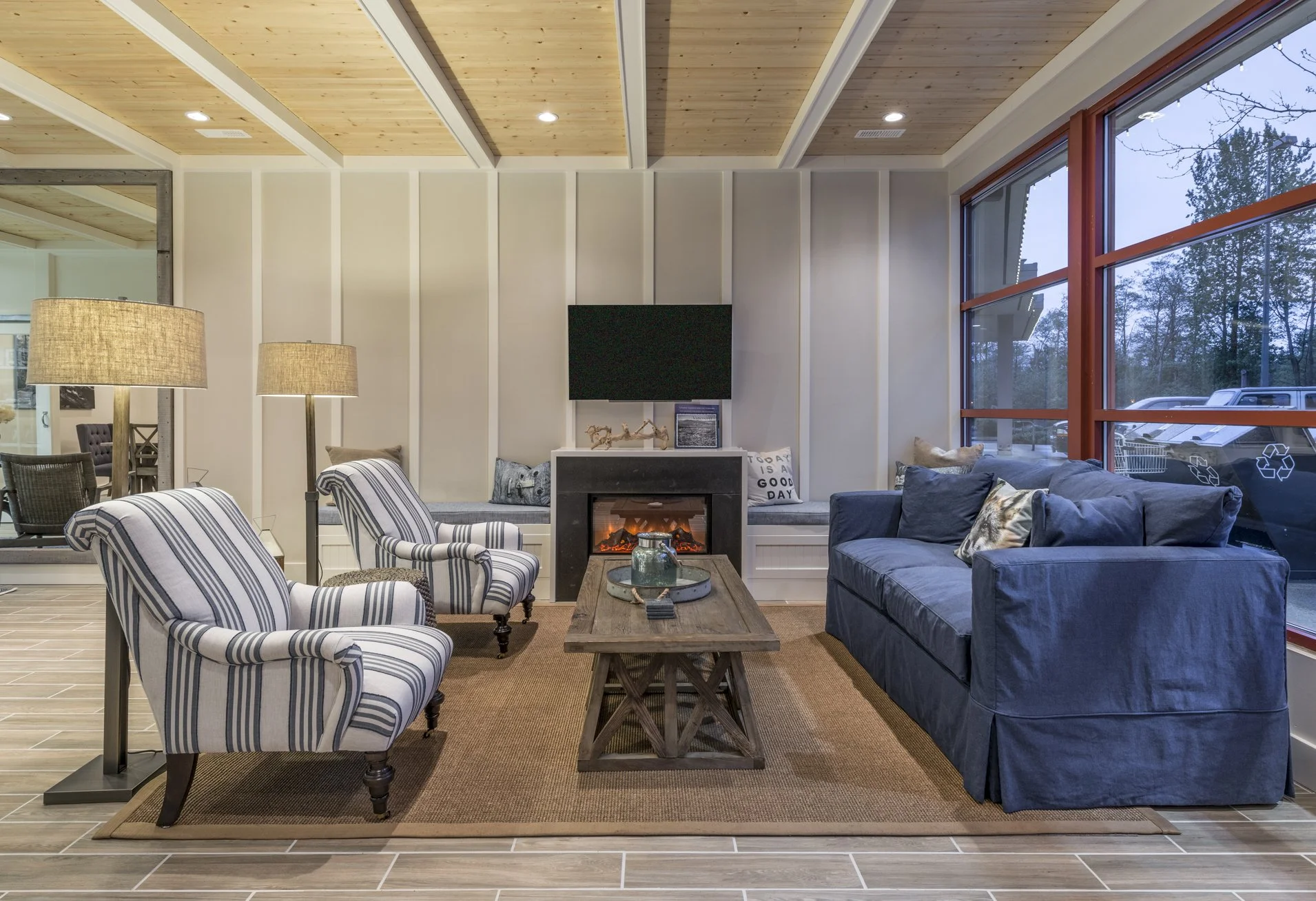 Living room with striped armchairs, a blue sofa, a wooden coffee table, a fireplace, a wall-mounted TV, and large windows showing trees outside.