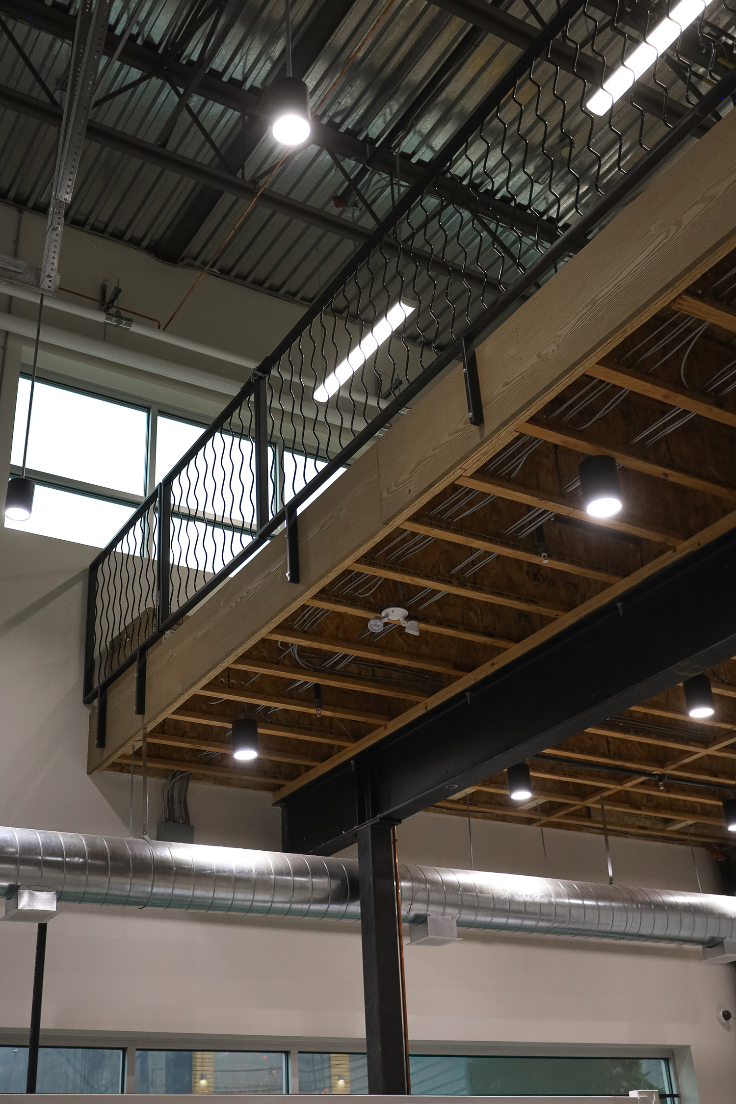 Interior view of a modern industrial-style building with exposed metal and wood beams, ductwork, and railing on a mezzanine level, illuminated by ceiling lights.