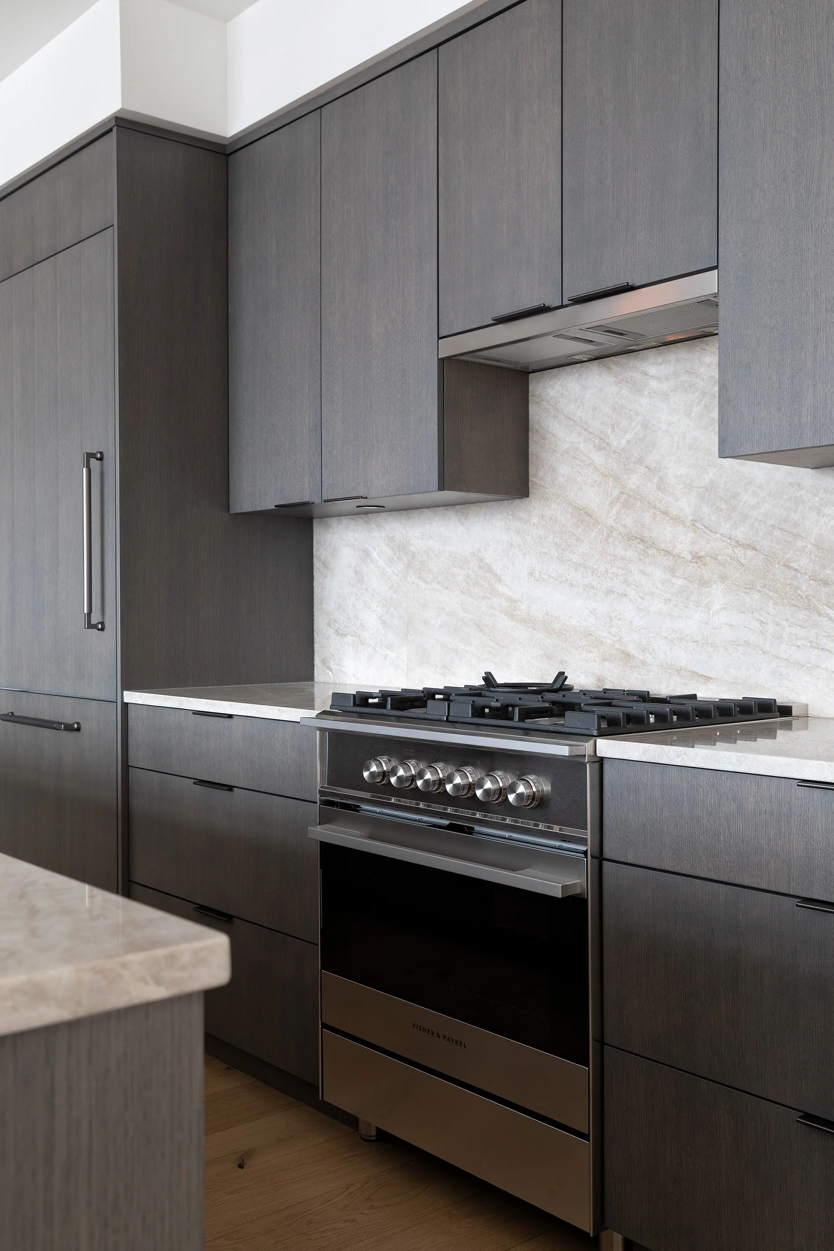 Modern kitchen with dark gray cabinets, stainless steel oven, and beige stone backsplash.