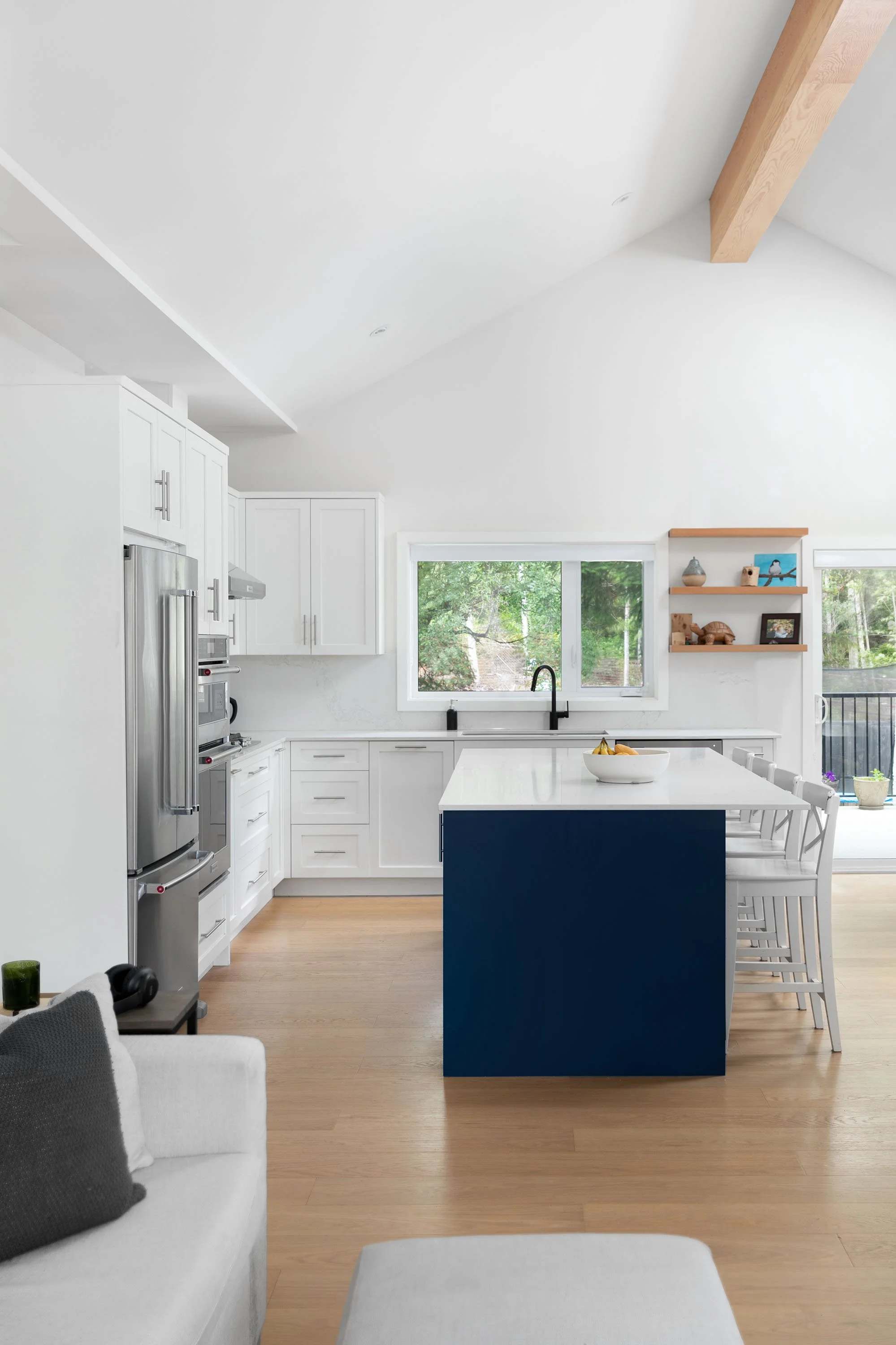 Modern kitchen with white cabinets, stainless steel refrigerator, window overlooking trees, and a central island with a blue base and white countertop. There are white chairs around the island and a small white bowl of fruit.
