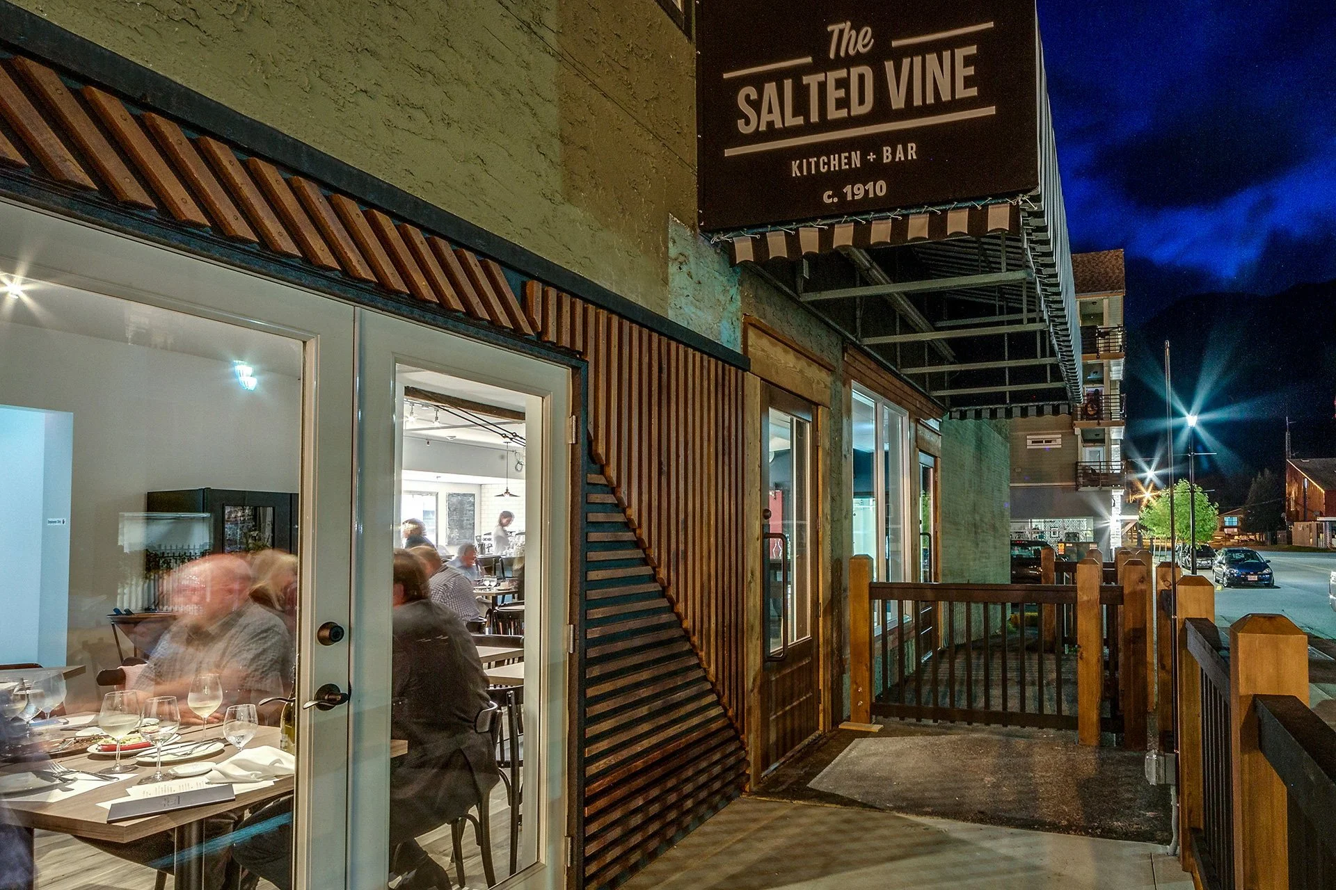 Nighttime view of the exterior of a restaurant called 'The Salted Vine Kitchen + Bar' with a patio area, through a glass door showing patrons dining inside. The building features wooden slats as exterior decor, and there is a parking lot with cars, streetlights, and mountains in the background.