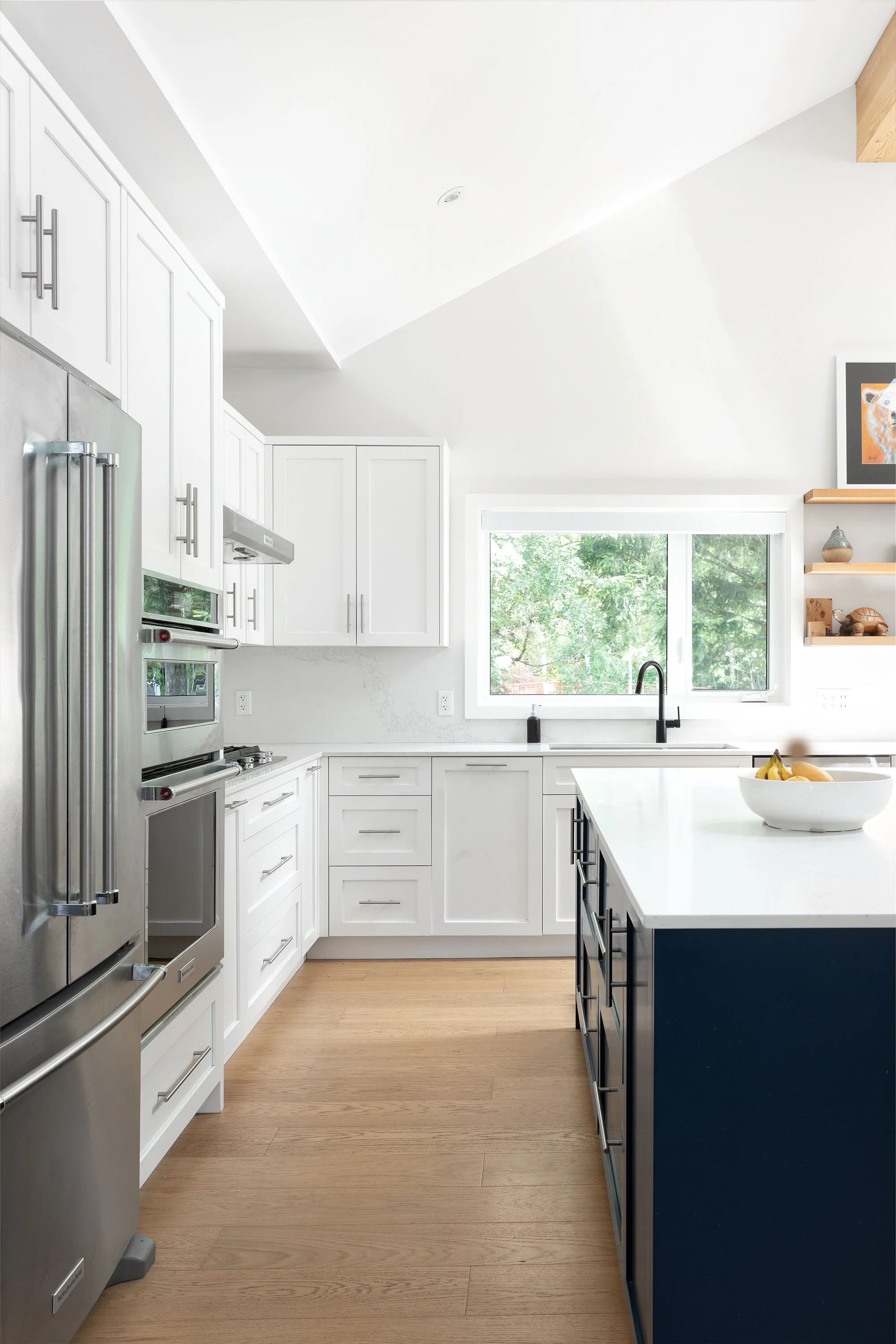 Modern kitchen with white cabinets, black island, stainless steel refrigerator, oven, and microwave. Light wood flooring, large window with greenery outside, and a bowl of bananas on the island.