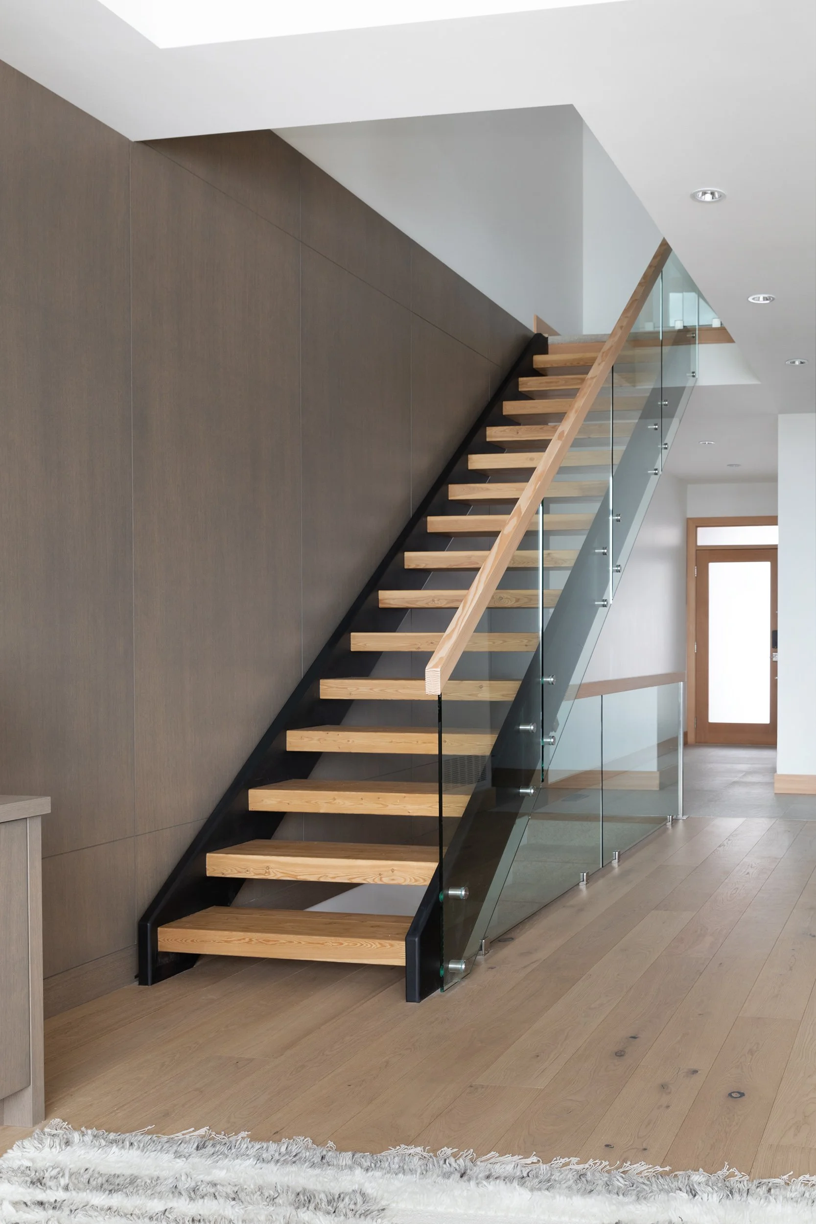 Modern staircase with wooden steps, glass railing, and black support frame in a minimalist interior with wood flooring and beige wall.