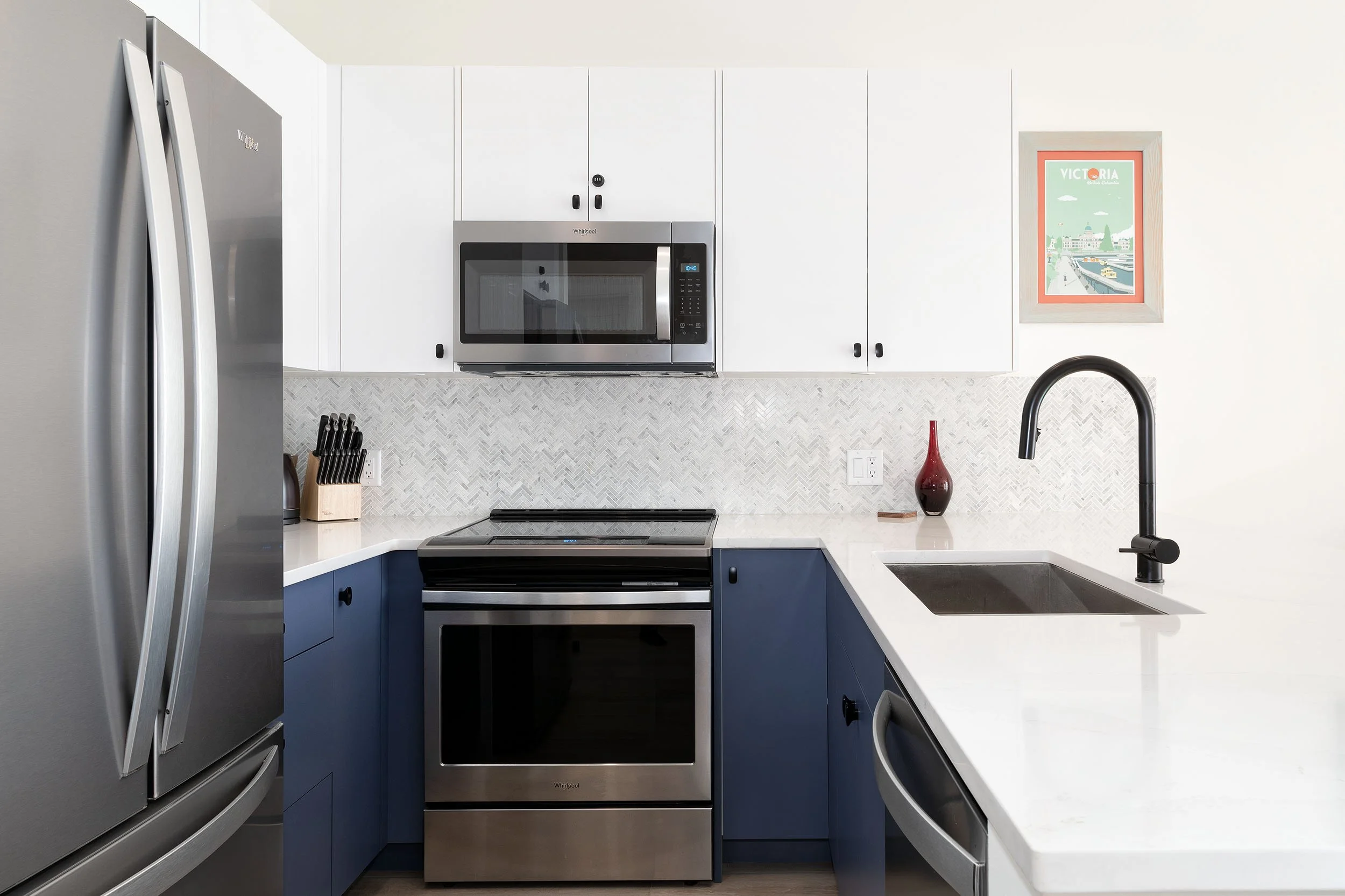 Modern kitchen with stainless steel refrigerator, microwave, and oven, navy blue lower cabinets, white countertop, and a framed poster on the wall.