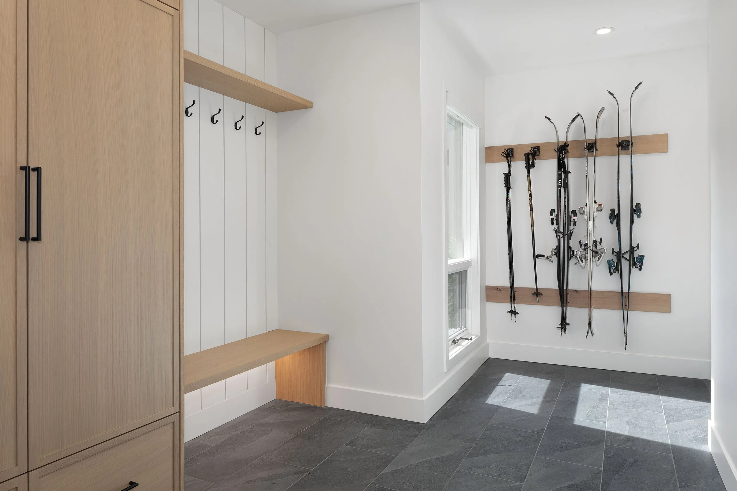 A modern entryway with a built-in wooden storage cabinet on the left, white paneled walls, black hooks on the wall, and a wooden rack holding several pairs of skis near a window.