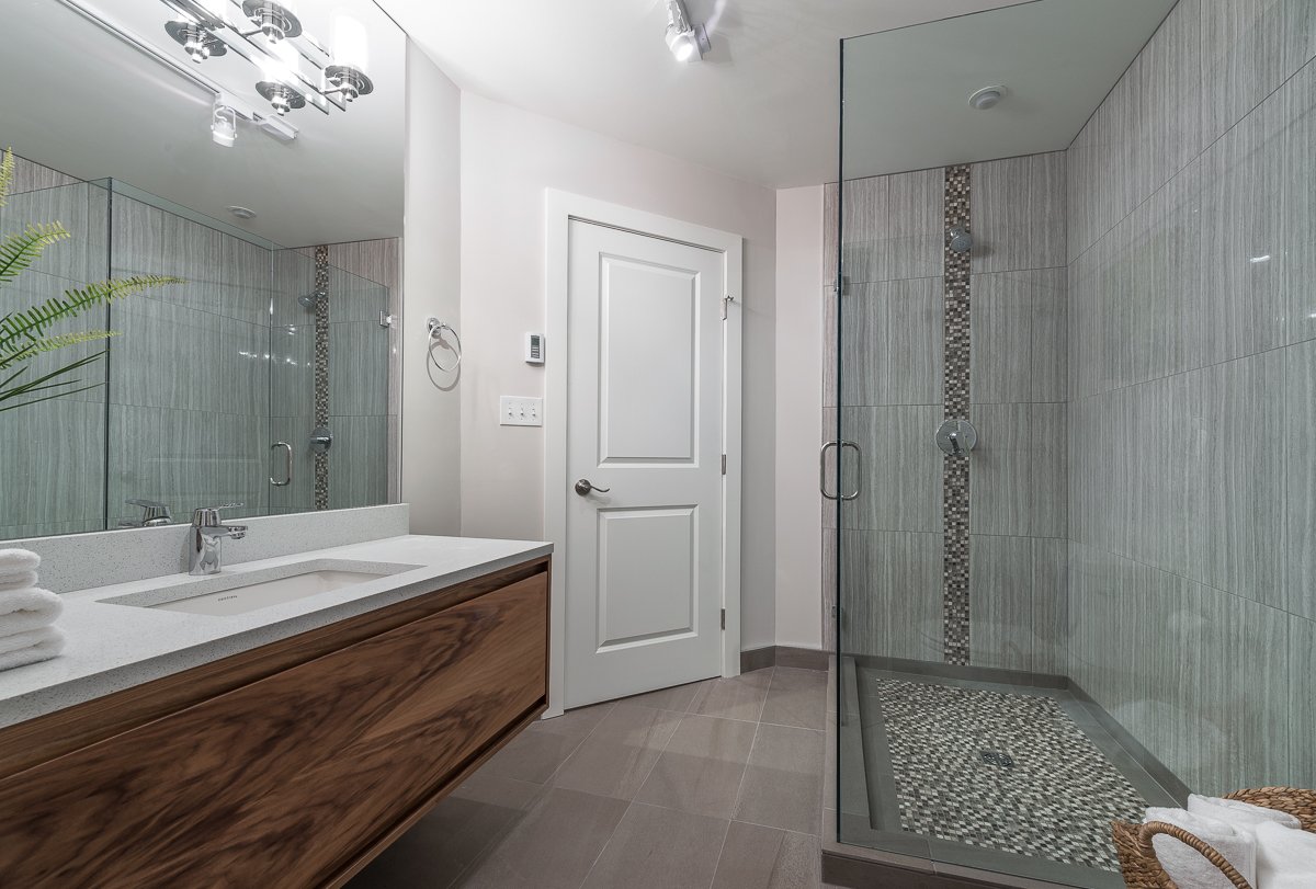 Modern bathroom with a large glass shower, wooden vanity with a white countertop, and a mirror with lighting fixtures above.