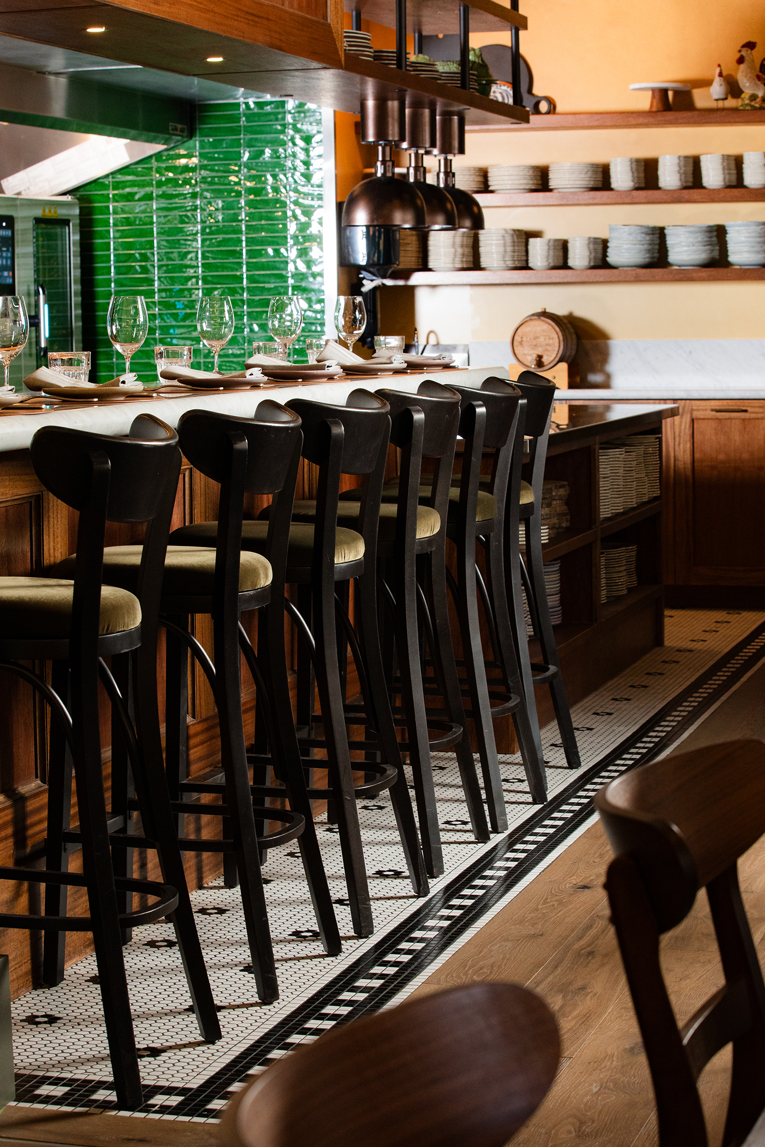 Interior of a restaurant with a bar counter set for dining, featuring wine glasses, plates, and utensils. Shelves with plates and decor are visible along with orange walls, green tiled wall, and black and white tiled flooring.