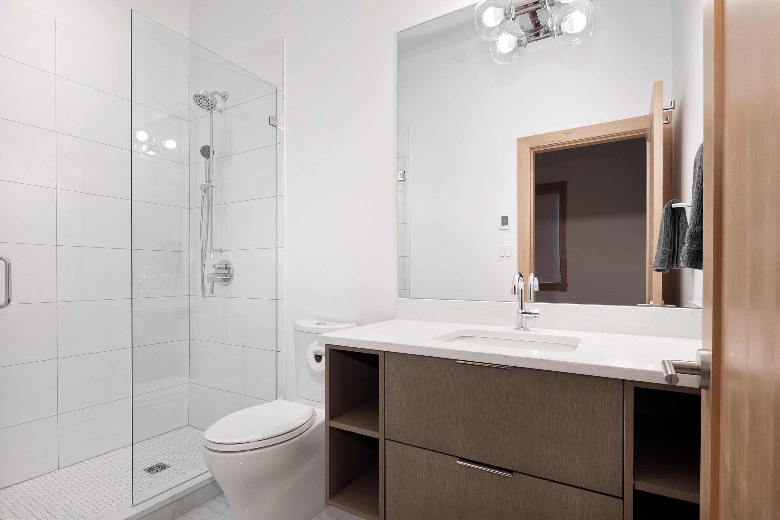 A modern bathroom with a glass-enclosed shower, a white toilet, a wooden vanity with a sink, a large mirror, and towels hanging on a wooden rack.