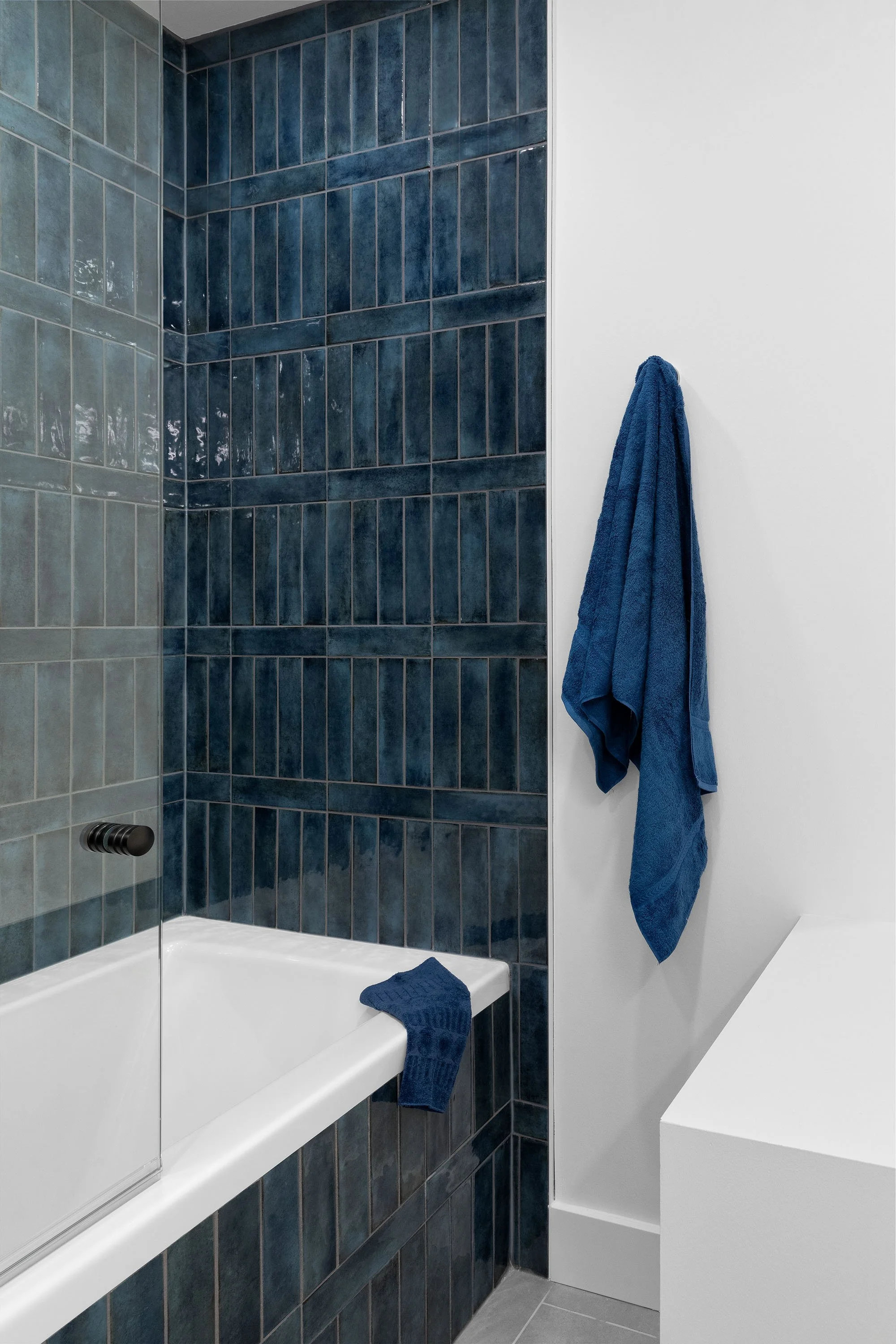 A modern bathroom shower area with dark blue tiles, a built-in shelf, a partial glass door, white walls, and blue towels hanging and placed on the edge of the bathtub.