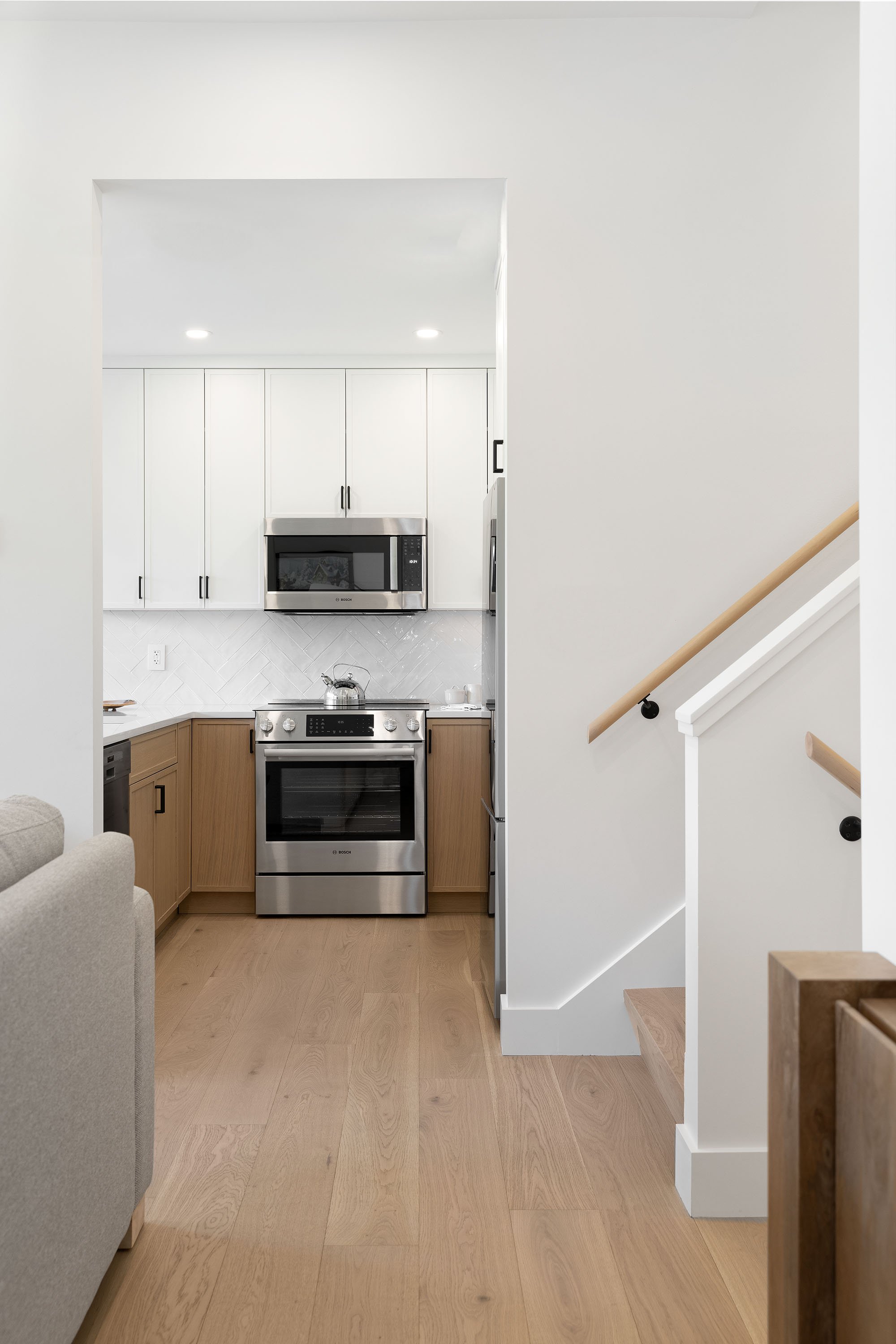 Interior view of a modern kitchen with white upper cabinets, stainless steel appliances including a microwave and oven, light wood lower cabinets, white walls, and light wood flooring.