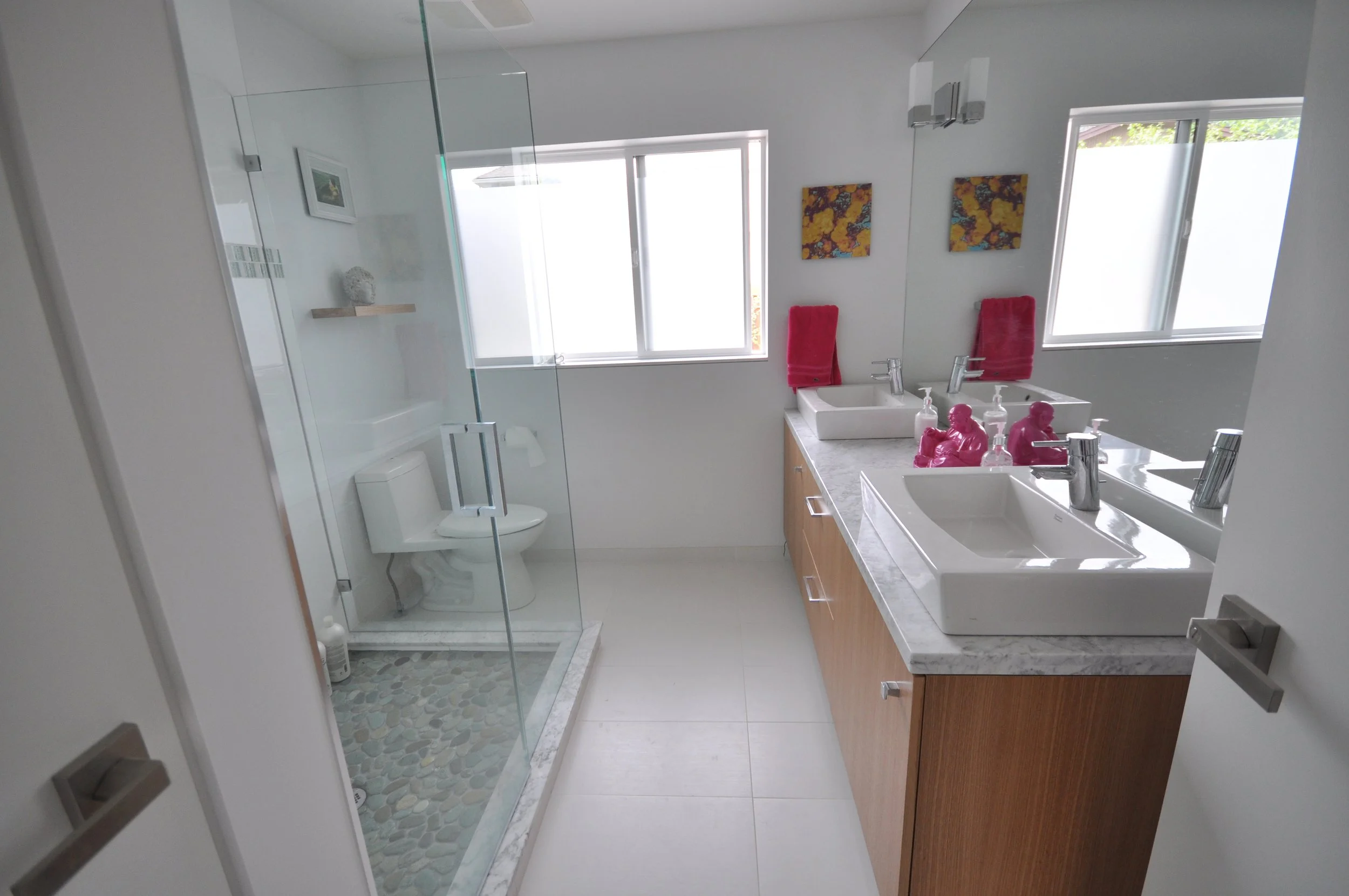 Modern bathroom with a glass-enclosed shower, white walls, a mirror, a double sink vanity with pink decorative items, red towels, and artwork on the wall.