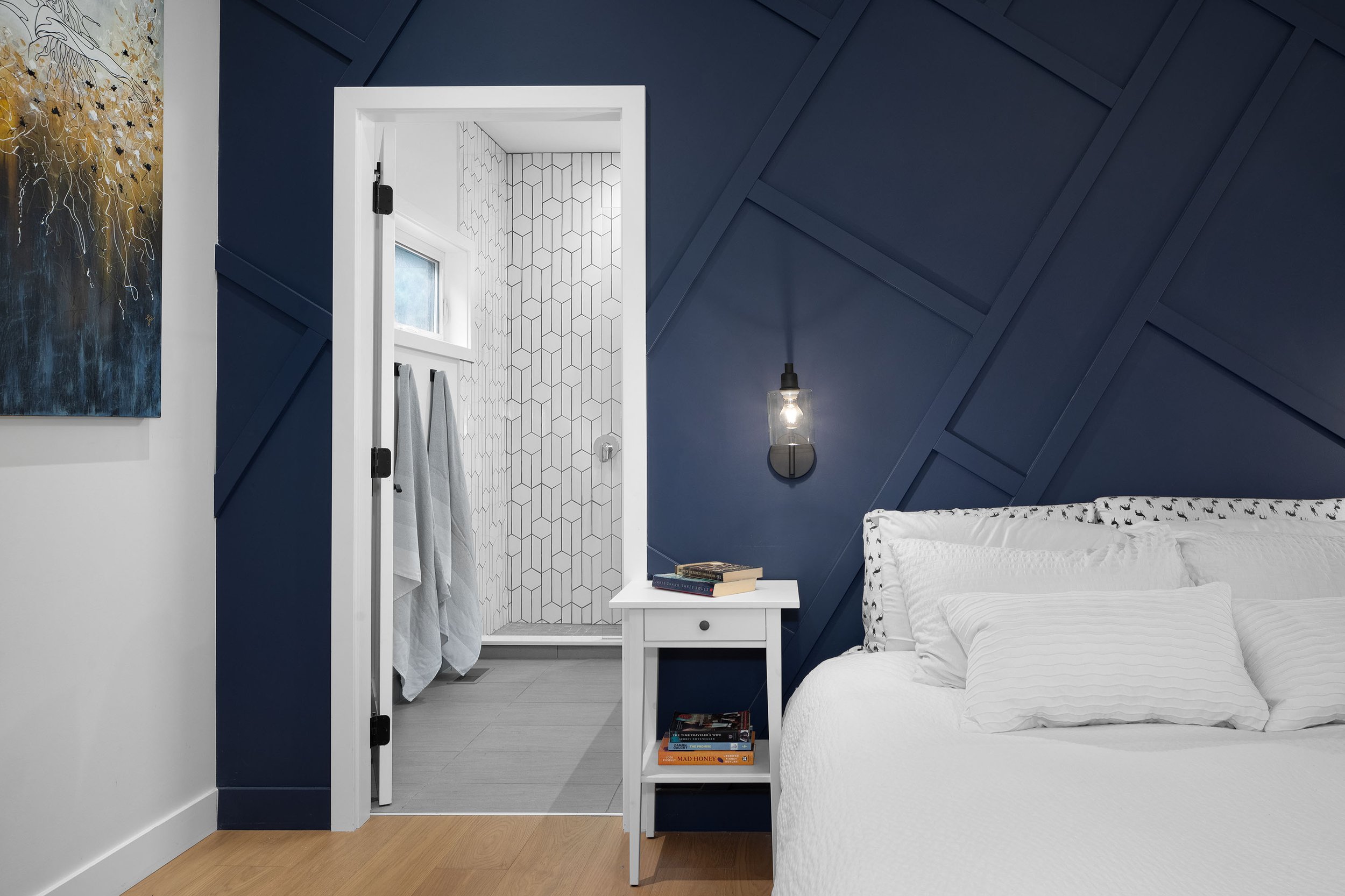 A bedroom with a white bed, blue wall, and windowed bathroom with patterned tiles through an open door.