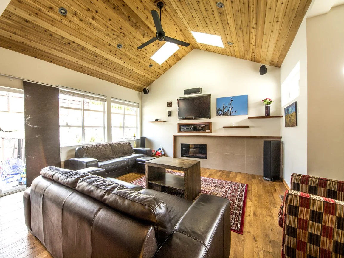 Living room with a vaulted wooden ceiling, leather sofas, a fireplace, a mounted television, windows with blinds, hardwood flooring, and decorative wall shelves.