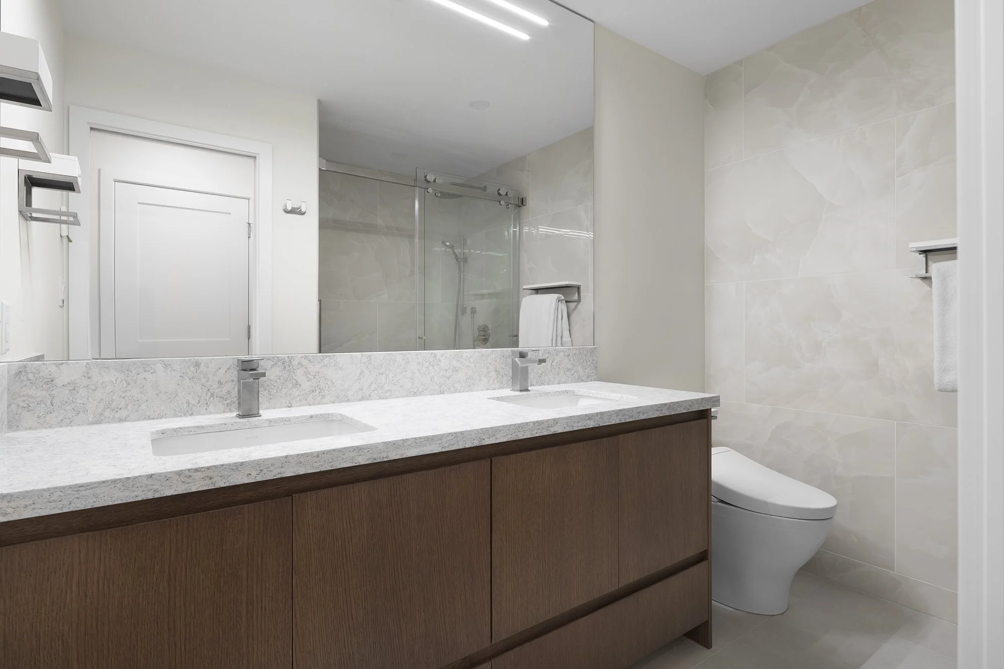 Modern bathroom with double vanity, marble countertop, wall-mounted faucet, large mirror, walk-in shower with glass door, beige tiled walls, and a wall-mounted toilet.