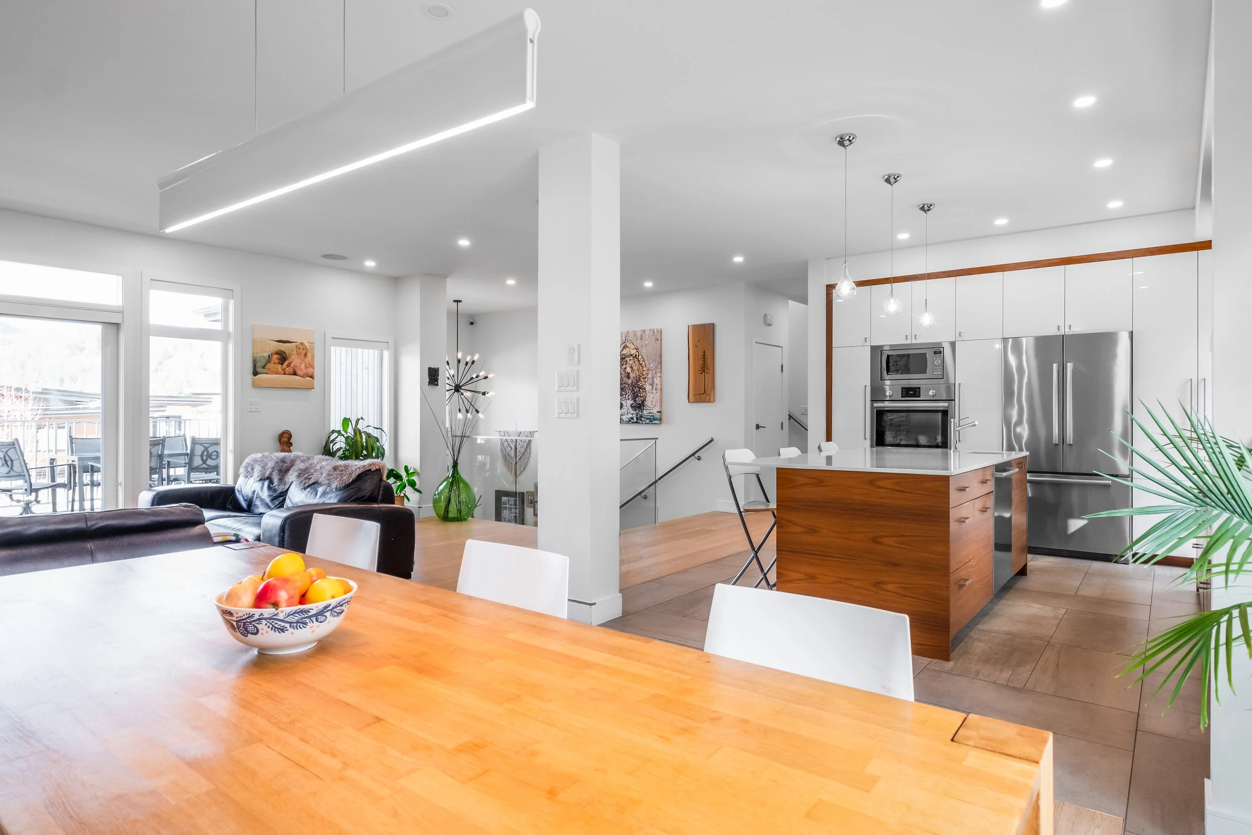 Open-concept modern kitchen and living room with white walls, wooden floors, stainless steel appliances, minimalistic furniture, and natural light from large windows.