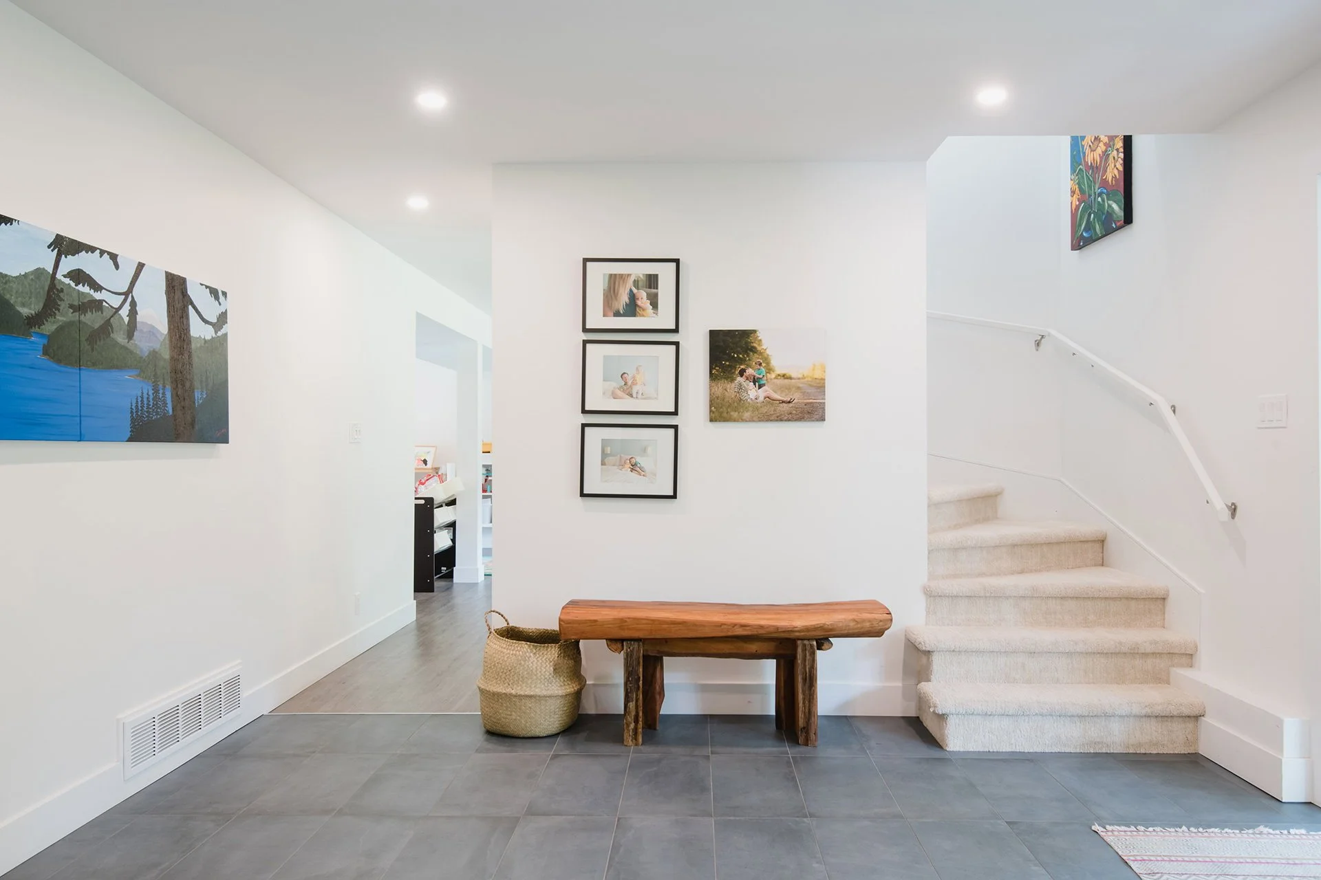 Interior view of a modern home entryway with white walls, framed family photos, a wooden bench, a woven basket, a set of carpet stairs, and minimalist lighting.