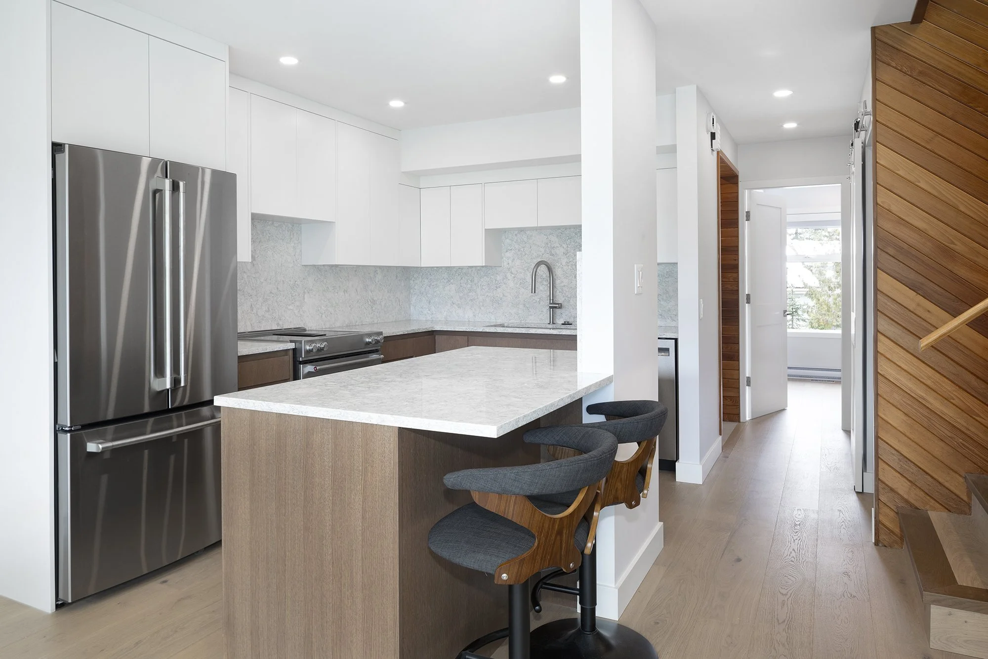 Modern kitchen with white upper cabinets, wooden lower cabinets, and stainless steel refrigerator, island with granite countertop, and bar stools, adjacent to a hallway with wood-paneled wall and stairs.