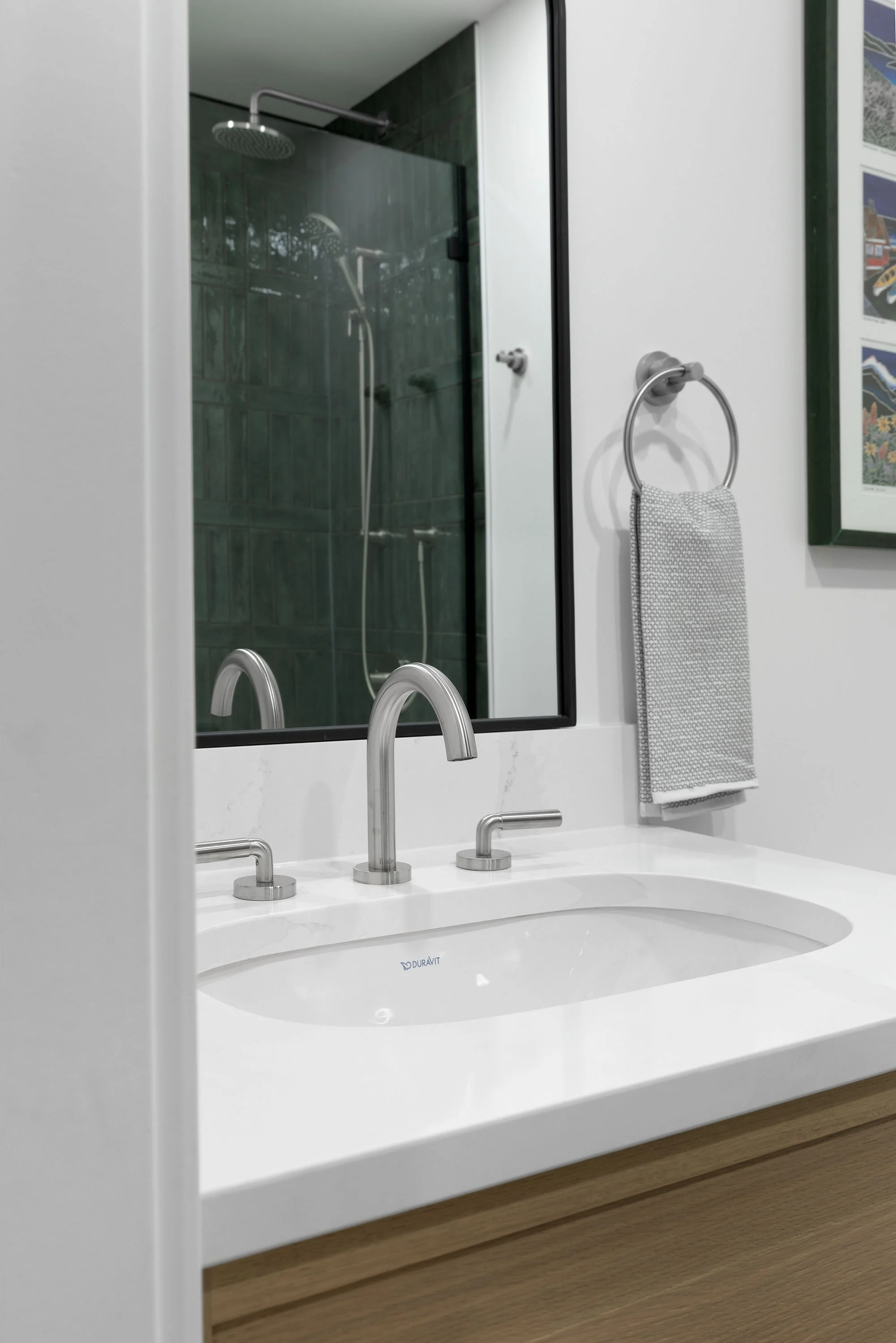 Modern bathroom with white vanity, double silver faucets, large mirror, and a walk-in shower with dark tile walls visible through the mirror.