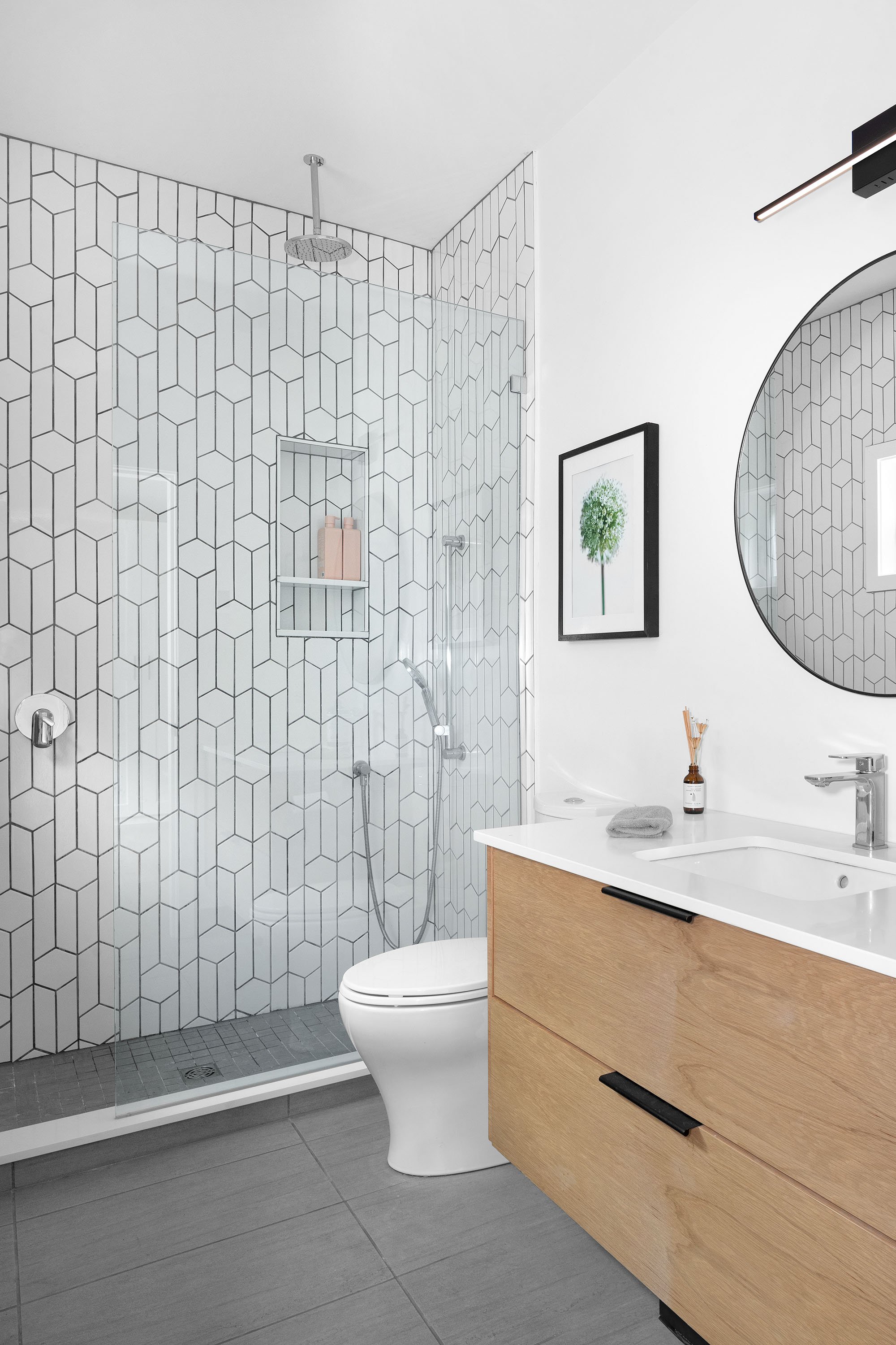 Modern bathroom with walk-in shower featuring geometric tile pattern, white wall, round mirror, wooden vanity with white sink, framed botanical art, and gray tile flooring.