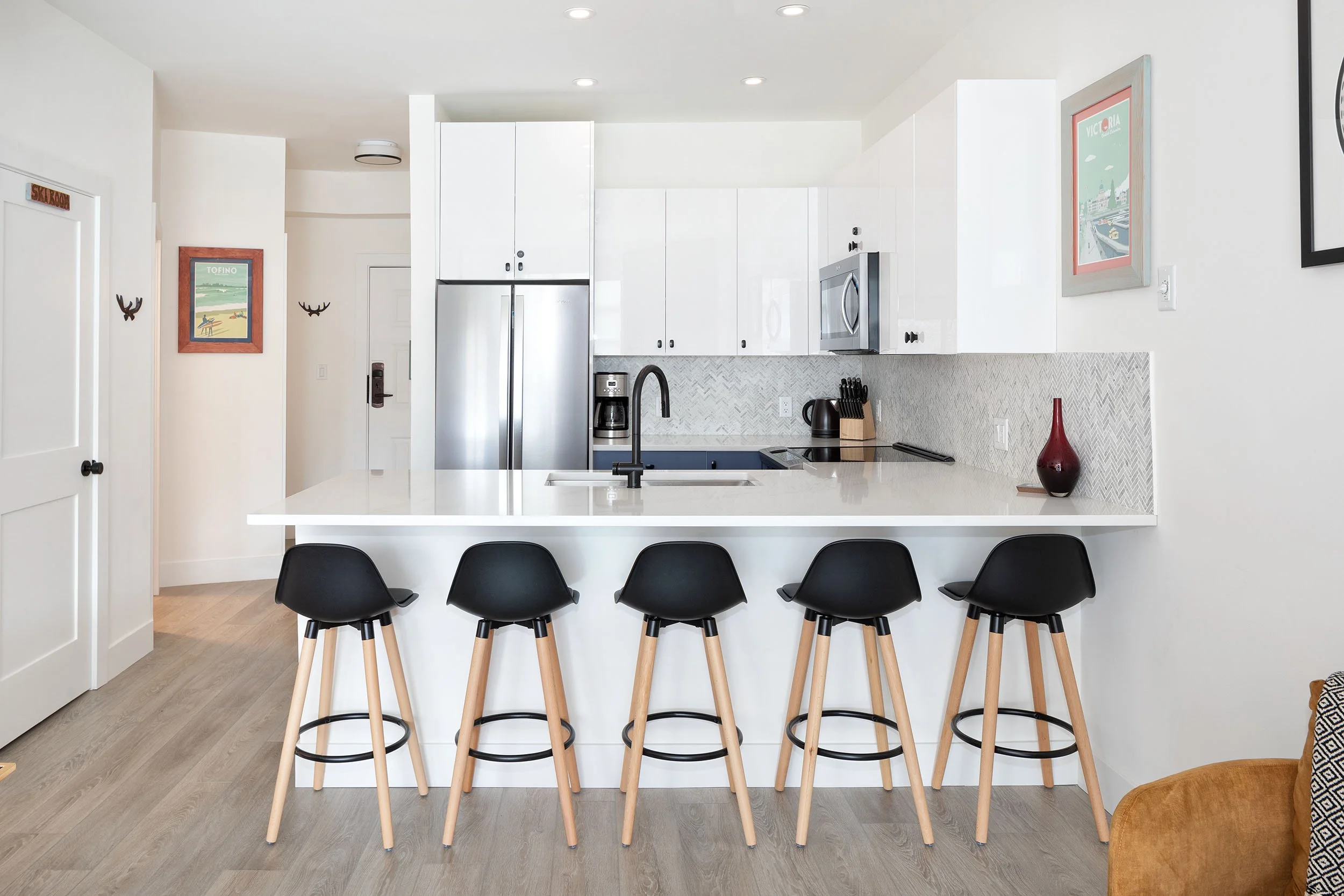 Modern kitchen with white cabinets, stainless steel refrigerator, coffee maker, electric kettle, black chairs around a white island, artwork on the walls, and wood flooring.