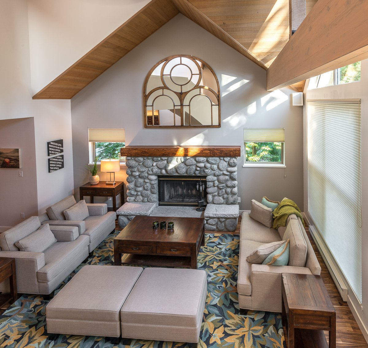 Living room with beige sofas, a wooden coffee table, stone fireplace, large windows with blinds, and a high vaulted ceiling with wood accents, sunlight streaming in.