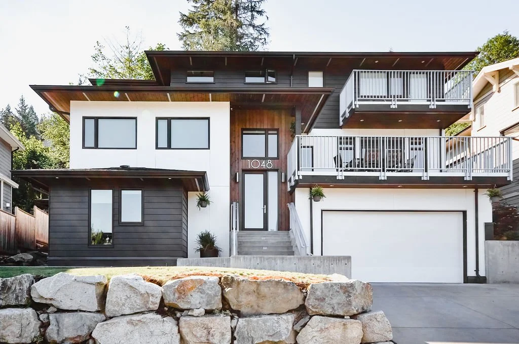 Modern multi-story house with white and dark gray exterior, large windows, two balconies with white railings, and a garage. Stone wall in front and greenery in background.