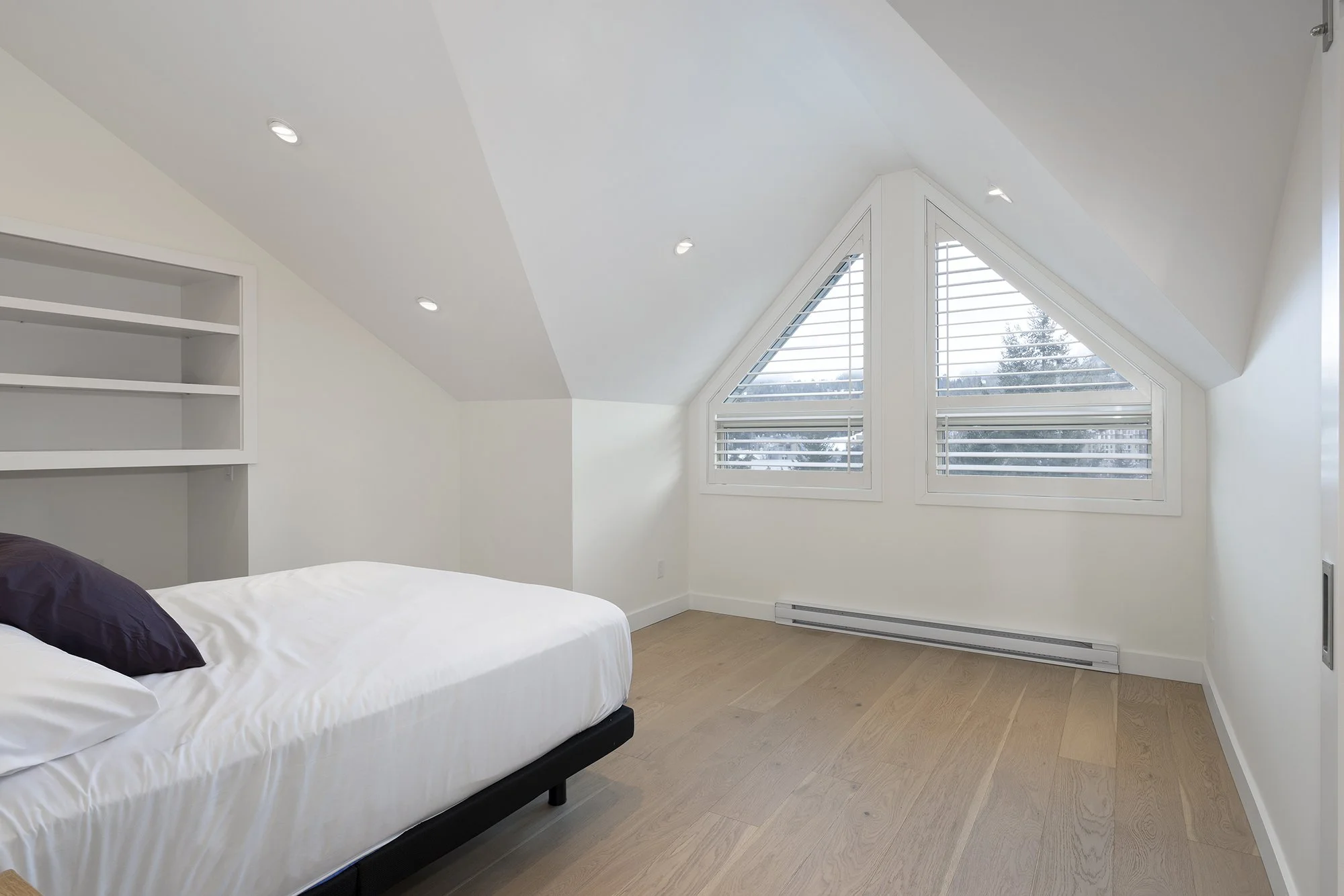 A bedroom with a sloped ceiling, large windows with shutters, a bed with white linens and purple pillows, and built-in shelves on the wall.