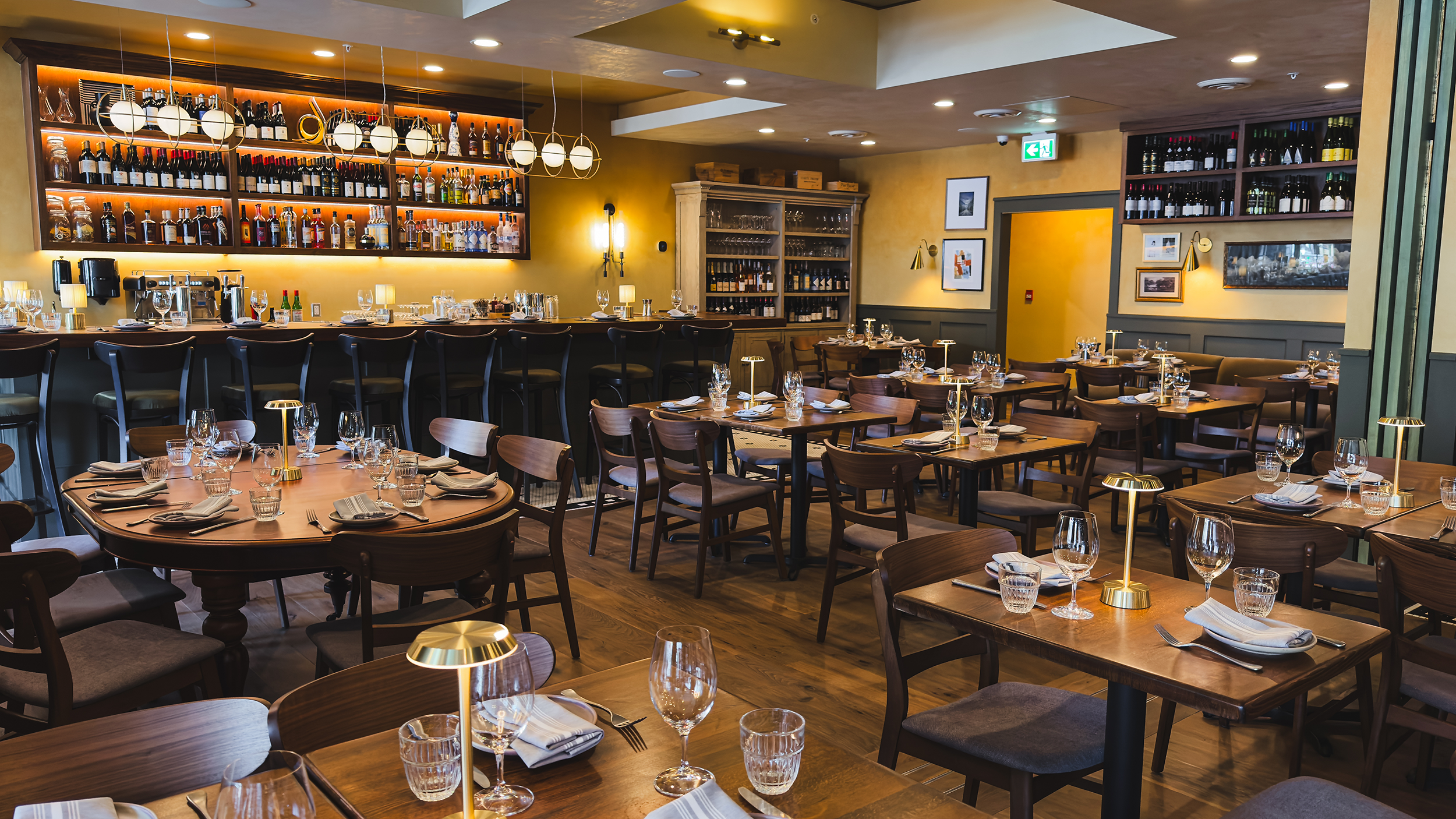 Interior of an elegant restaurant with wooden tables set with wine glasses, water glasses, napkins, and cutlery, illuminated by warm lighting, with shelves of wine bottles on the back wall.