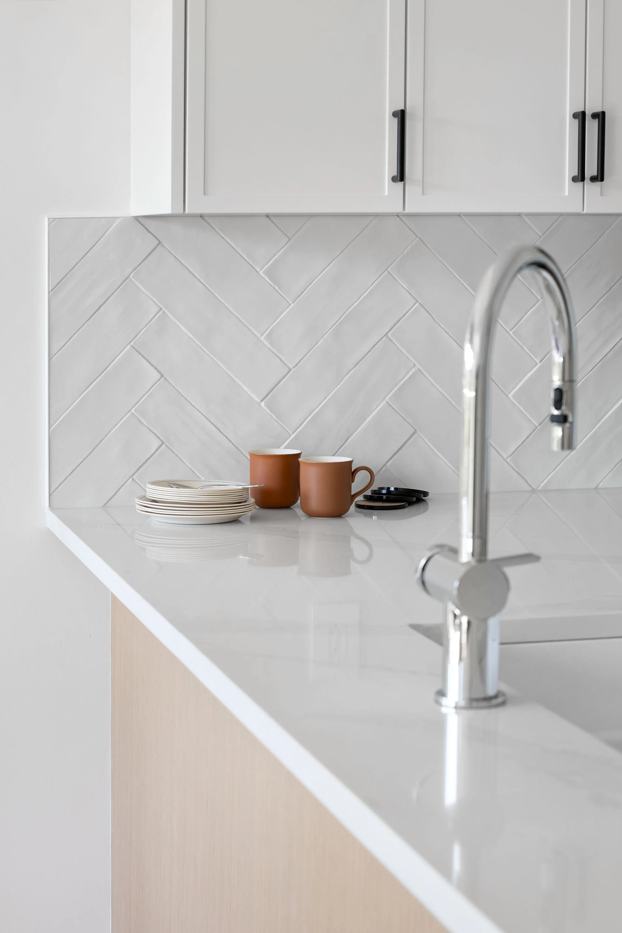 Modern kitchen with white cabinets, a white countertop, a chrome faucet, and a white tiled backsplash in a herringbone pattern. There are brown mugs, white plates, and black coasters on the counter.
