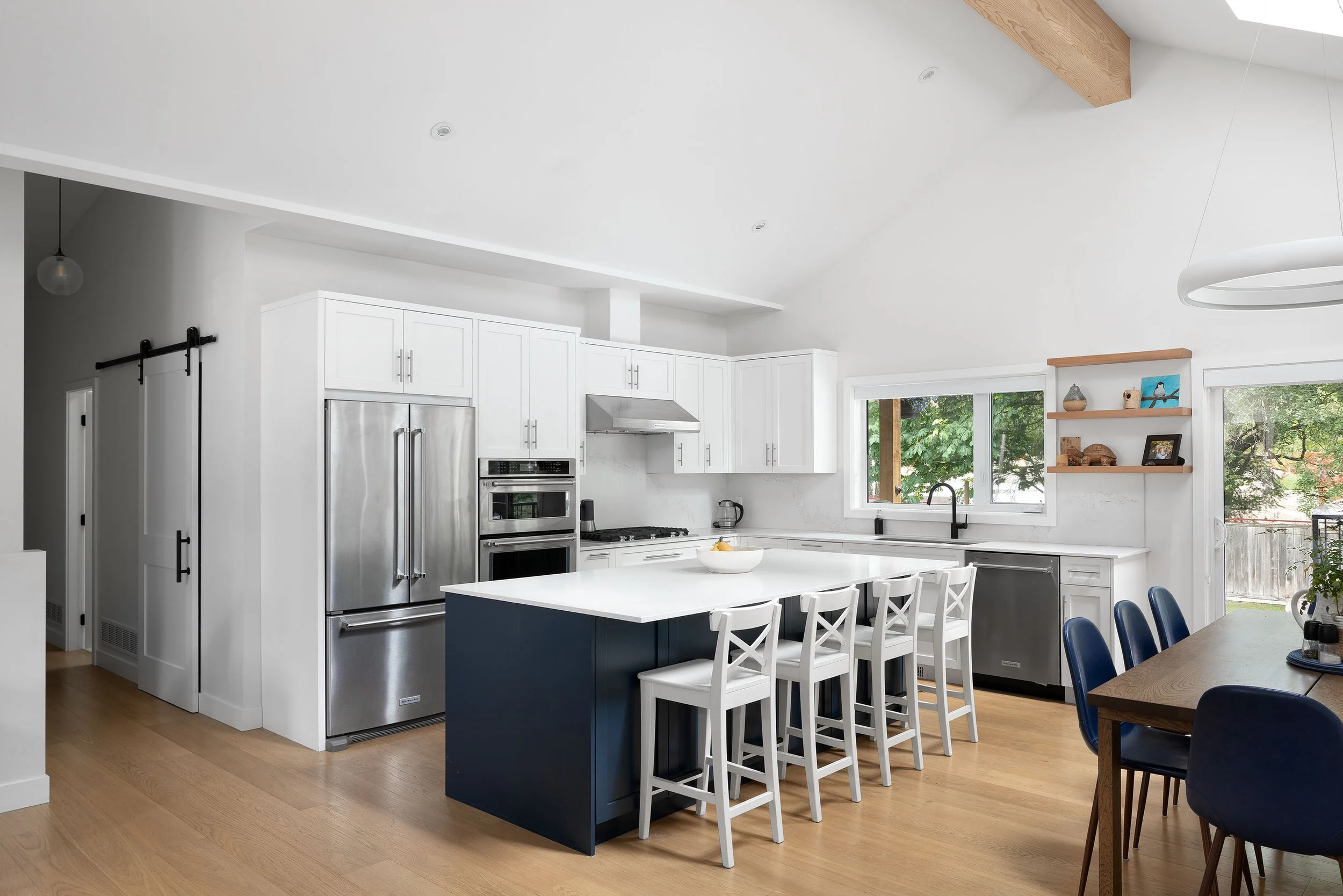 Modern kitchen with white cabinets, stainless steel appliances, and a white island with white chairs. Large window with a view of trees outside, and wooden flooring.