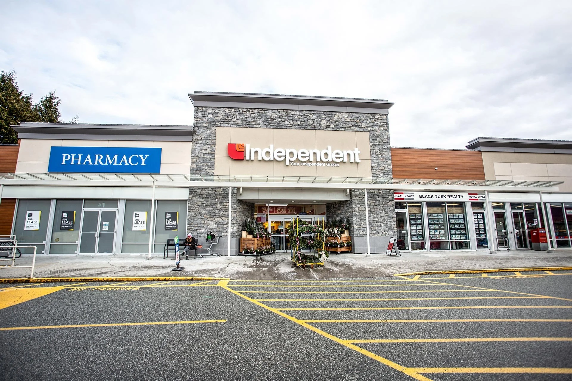 Exterior of a shopping center with a pharmacy, grocery store, and real estate office. There are parked shopping carts, a person sitting on a bench, and flowers for sale outside the stores. Overcast sky.