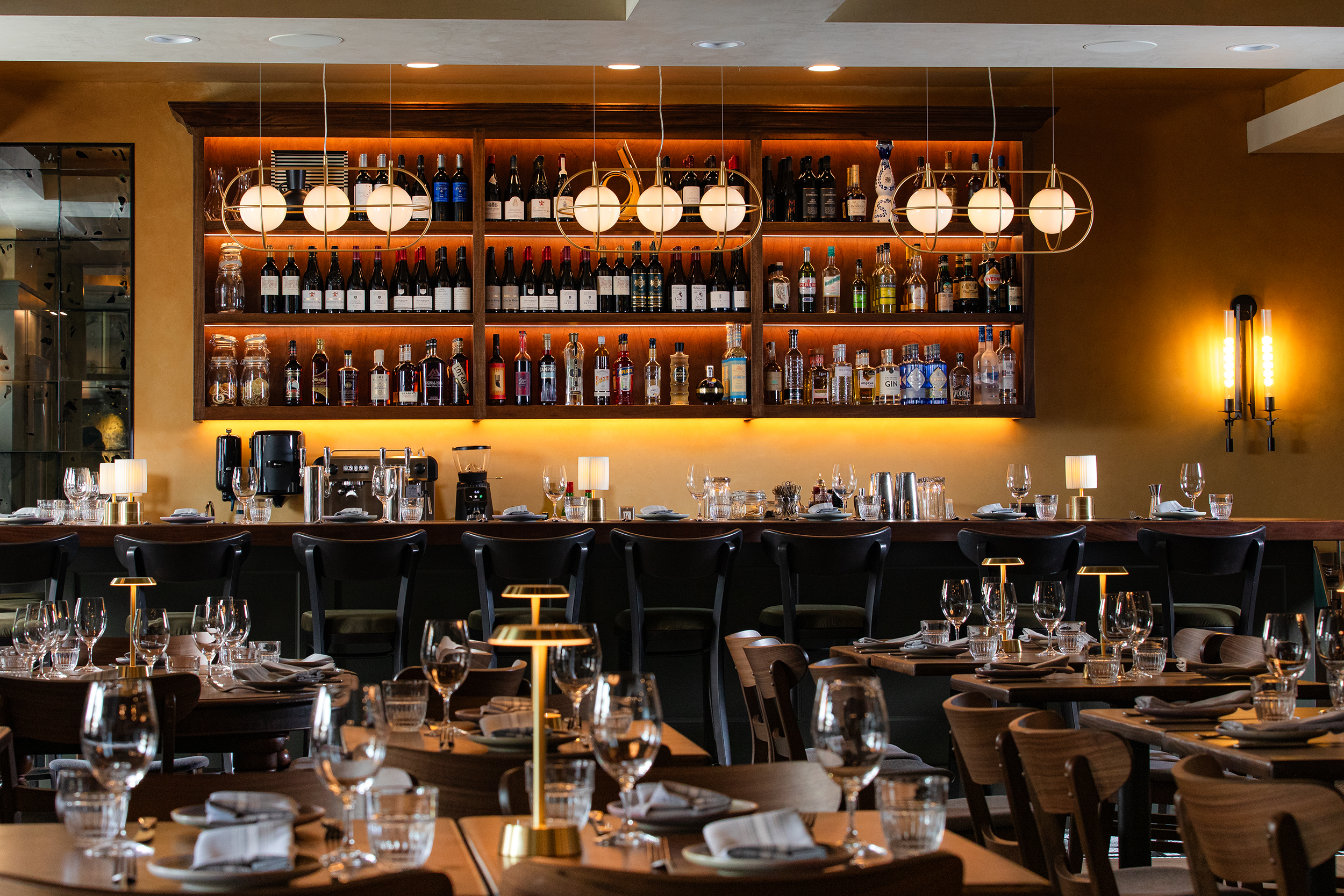 Interior of a restaurant with a bar in the background, shelves stocked with wine and spirits, round modern pendant lights, and a dining area with tables set with glasses and napkins.