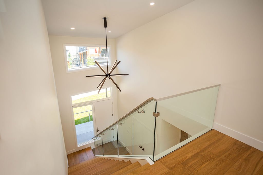 Interior view of a modern residential staircase with wooden steps and glass railing, leading down to a front door with large window, in a bright, white-walled space with a contemporary ceiling fan.