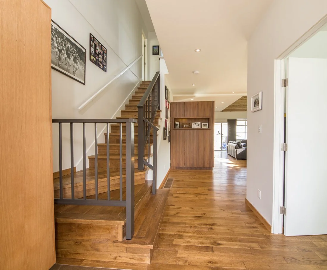 Interior view of a home with wooden floors, stairs with a metal railing, and a partial view of a living room with couch and large windows in the background.