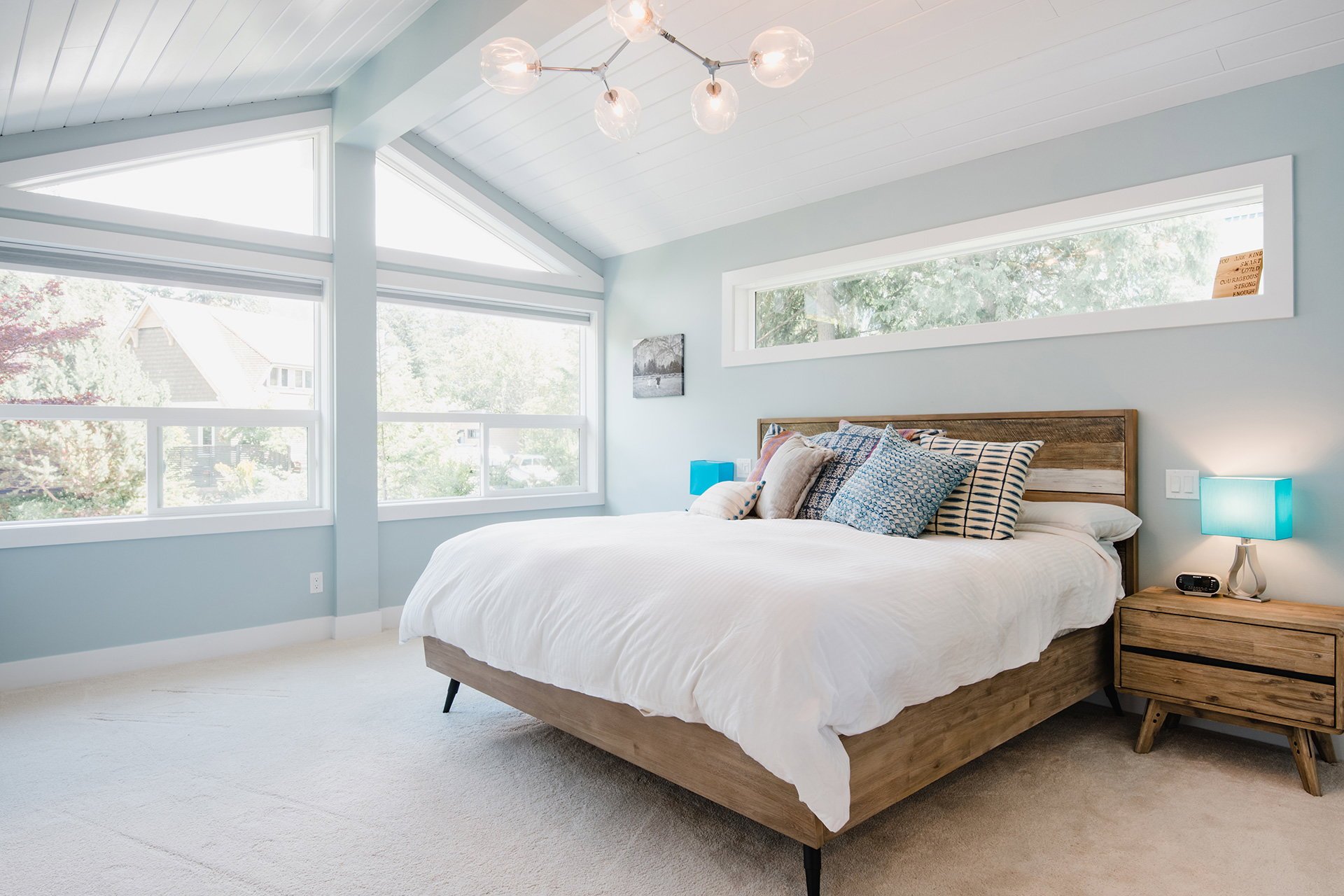 A bedroom with white bedding and multiple pillows on a wooden bed frame. Large windows providing natural light, a modern chandelier, a nightstand with a blue lamp, and a small picture on the wall.