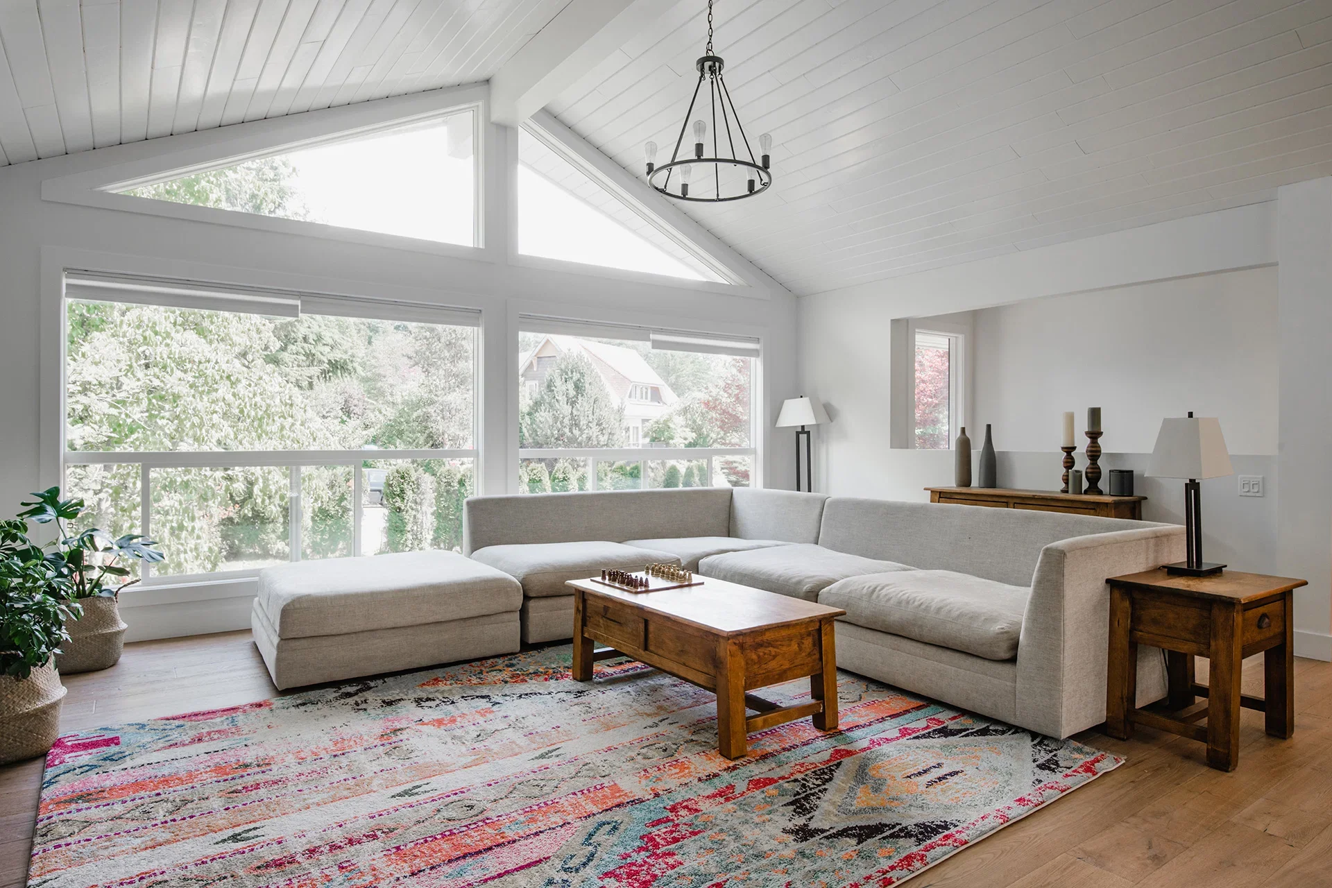 Living room with large windows, white walls, and a vaulted ceiling with a black chandelier. A beige sectional sofa faces a wooden coffee table, with a colorful rug underneath. There are side tables with lamps and decorative vases, and a potted plant 