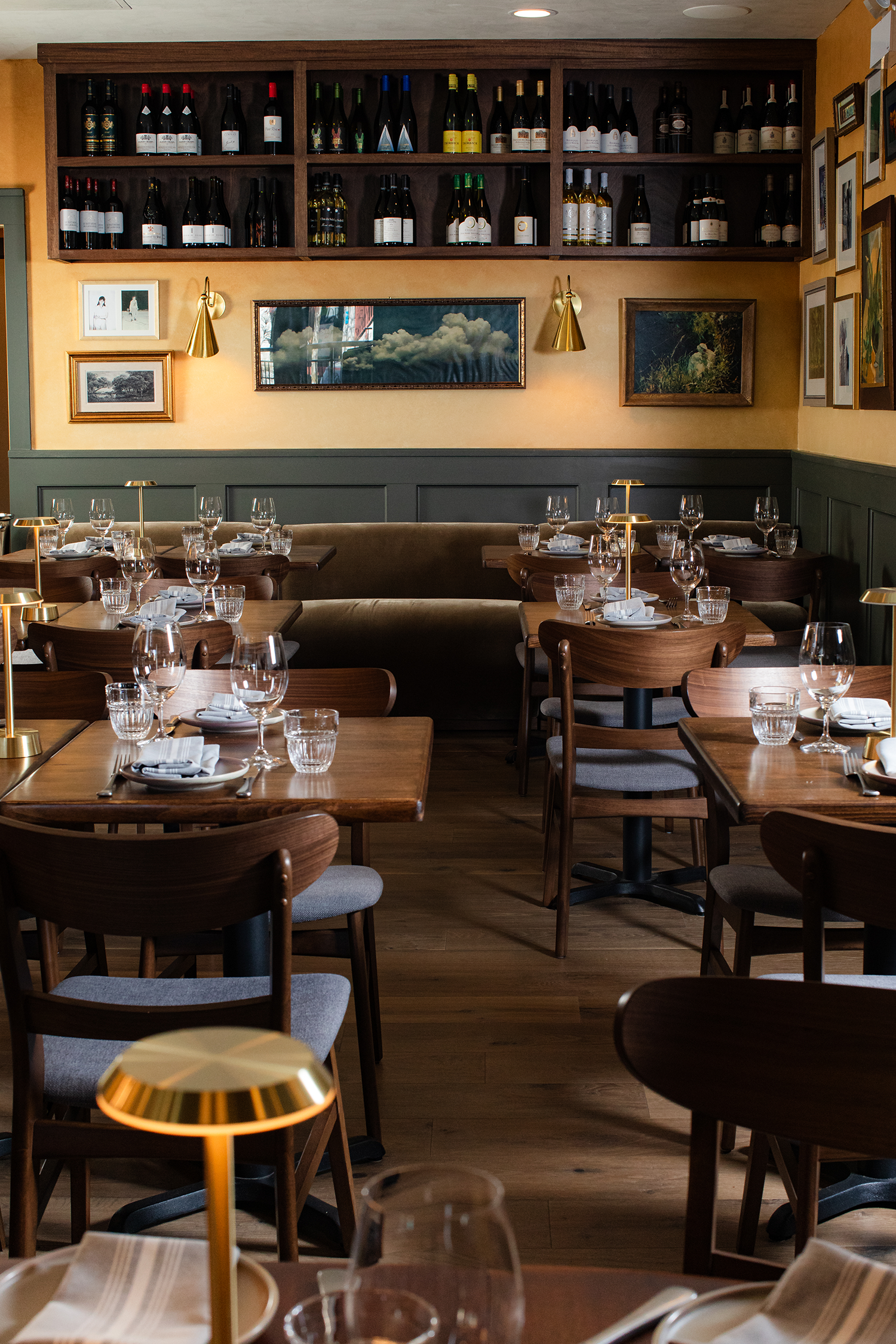 A restaurant dining room with wooden tables set with glasses, napkins, and plates, gold-colored lamps on each table, and a wall decorated with framed artwork and a large shelf with various bottles of wine.