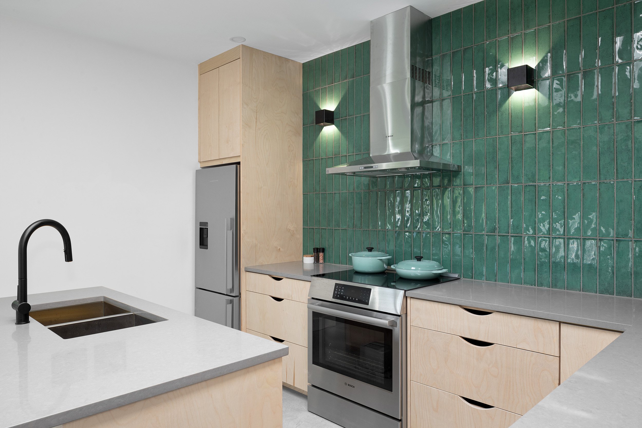 Modern kitchen with light wood cabinets, gray countertops, green tiled backsplash, stainless steel appliances, black faucet, and two black wall-mounted lights.