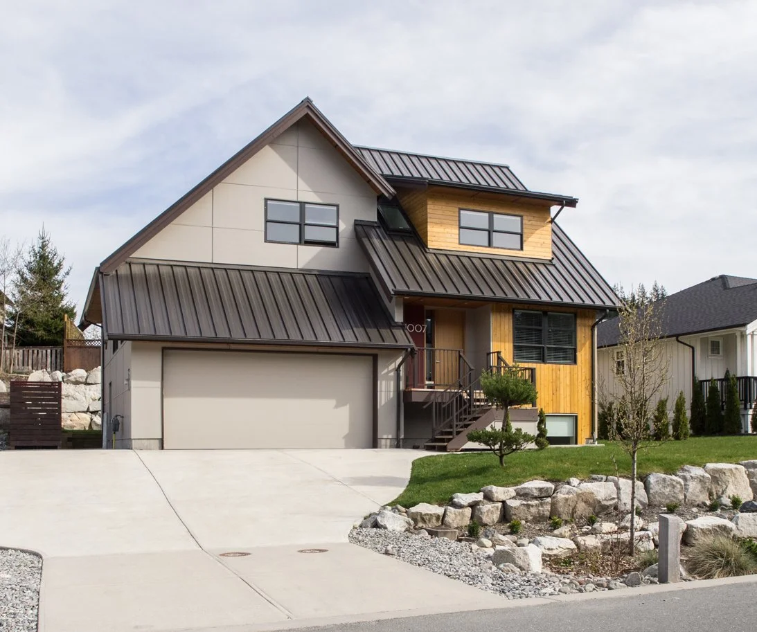 Modern two-story house with a gray metal roof, a two-car garage, and a small front yard with rocks and trees.