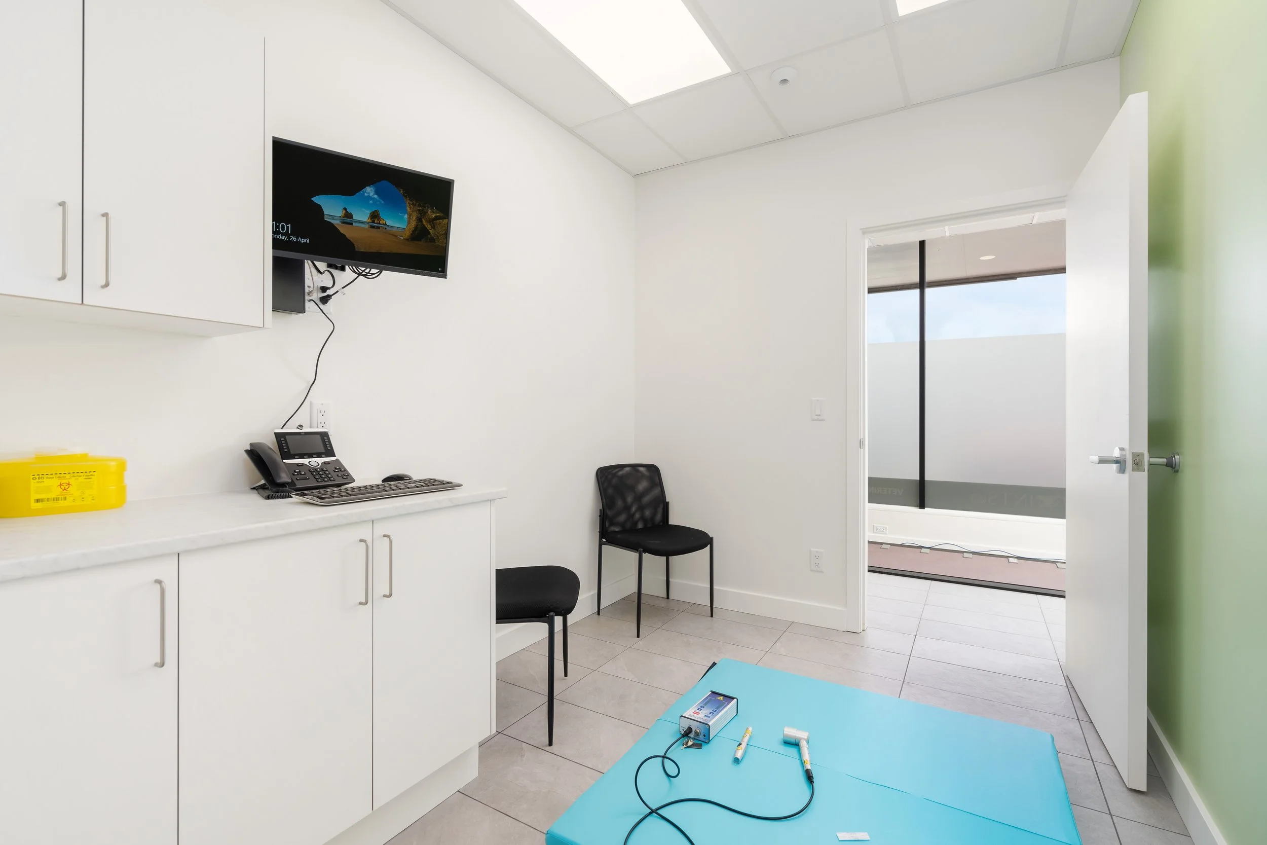 Medical examination room with white cabinets, a desktop computer, a wall-mounted monitor showing a beach scene, two black chairs, and medical supplies on a blue examination table.
