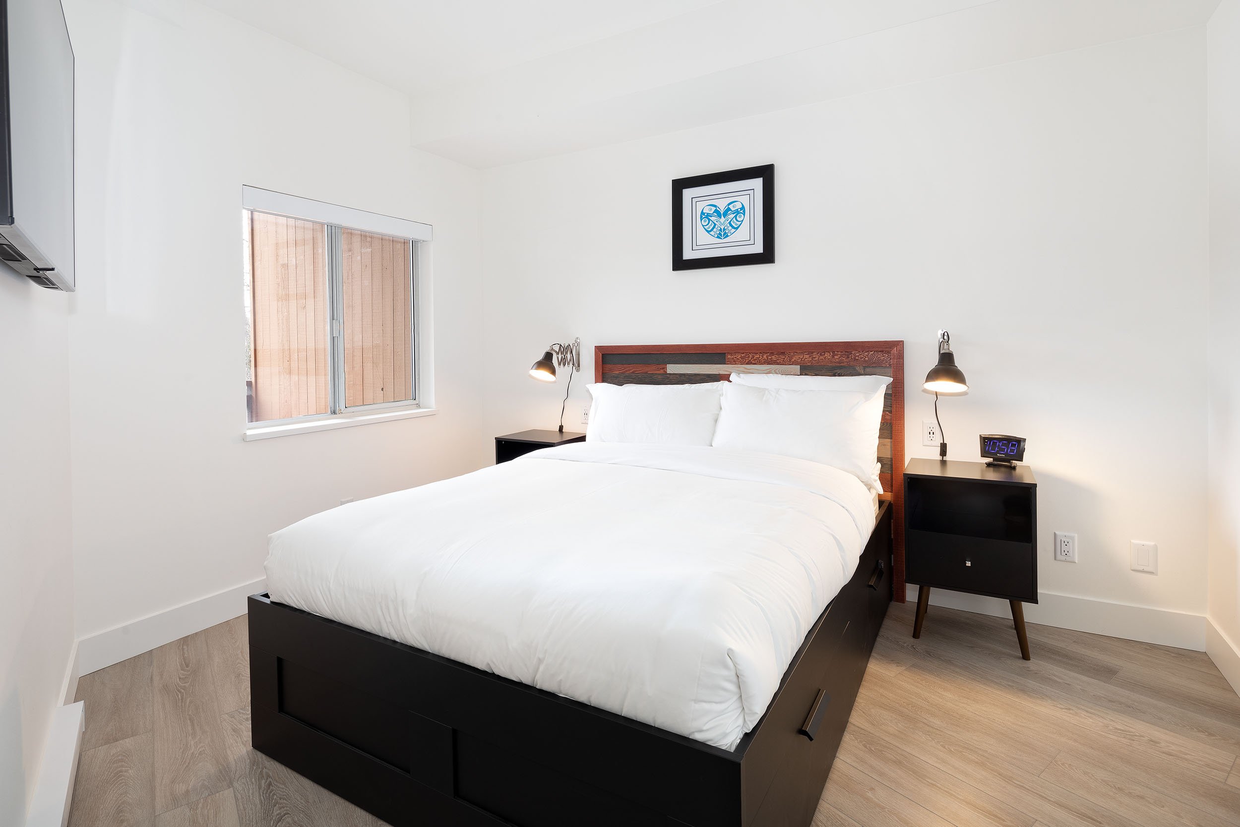 A modern bedroom with a white bed, black nightstands, and black and wood headboard. There are two black lamps on each side of the bed, a digital clock on the right nightstand, a window with blinds on the left wall, and a framed heart-shaped artwork o