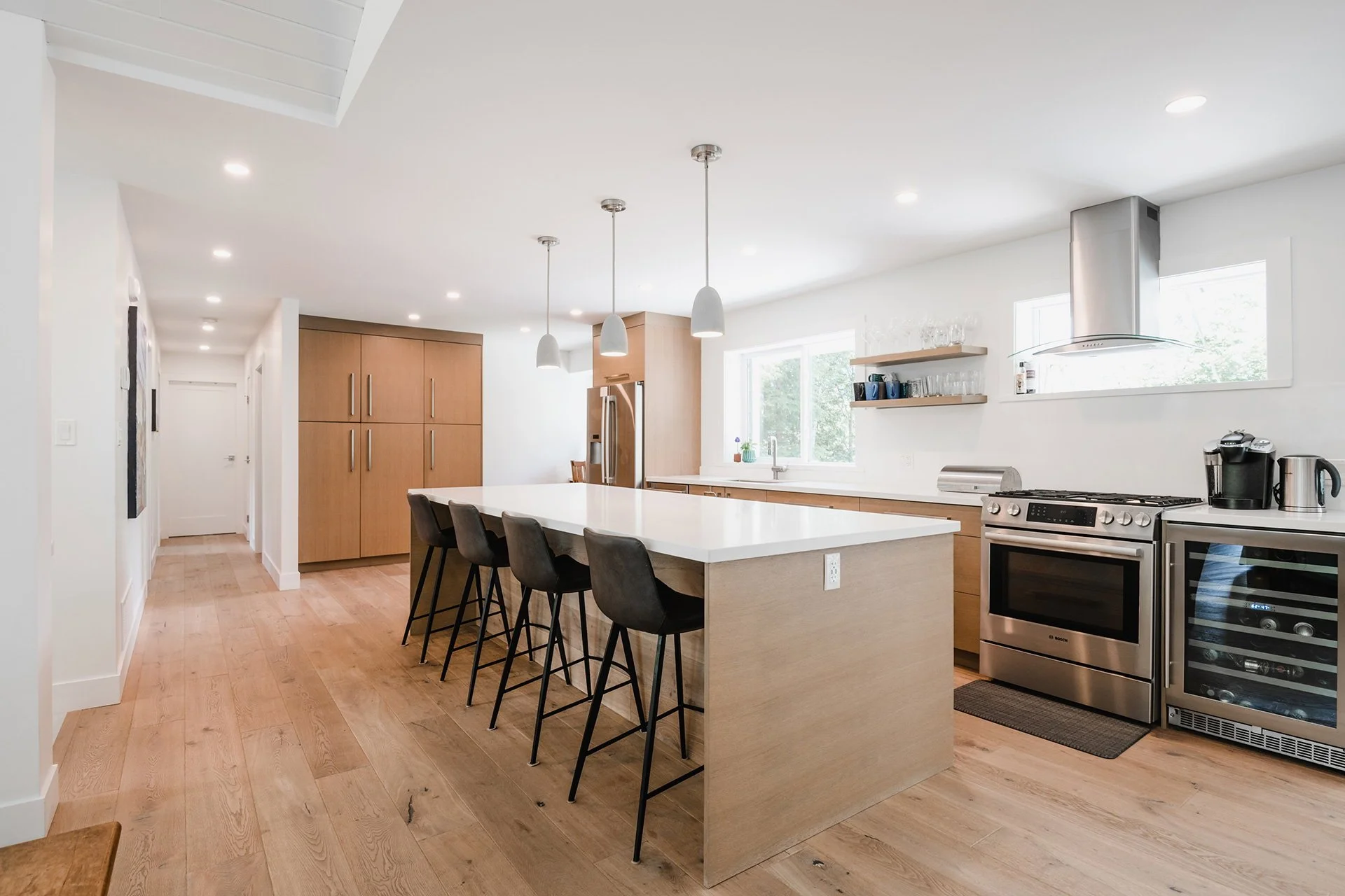 Modern kitchen with white countertops, stainless steel appliances, wooden cabinets, and black chairs around a kitchen island, illuminated by pendant lights.